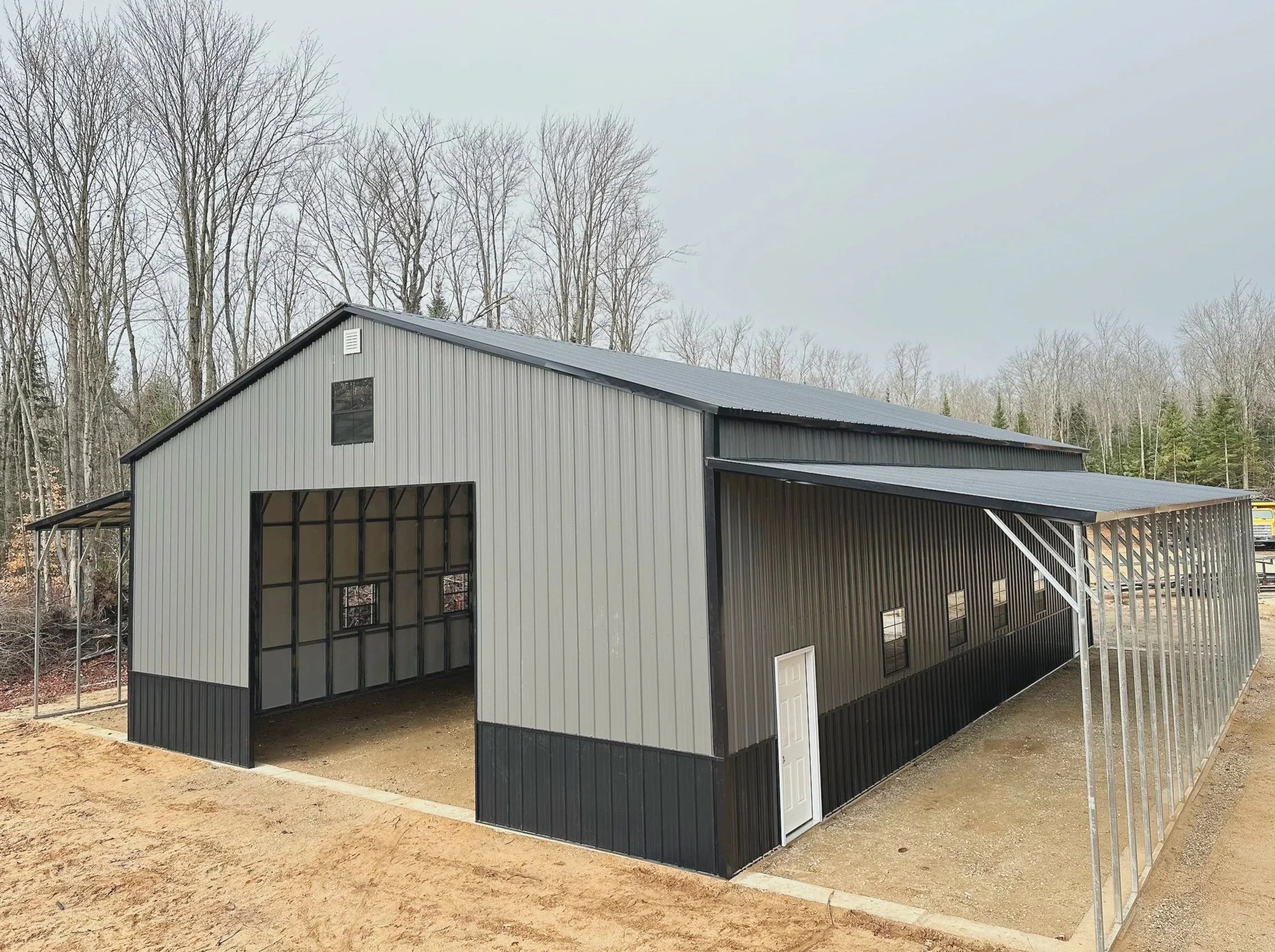 Large metal building under construction with black and gray exterior, multiple windows, and an open side revealing a spacious interior, situated in a rural area with trees in the background.