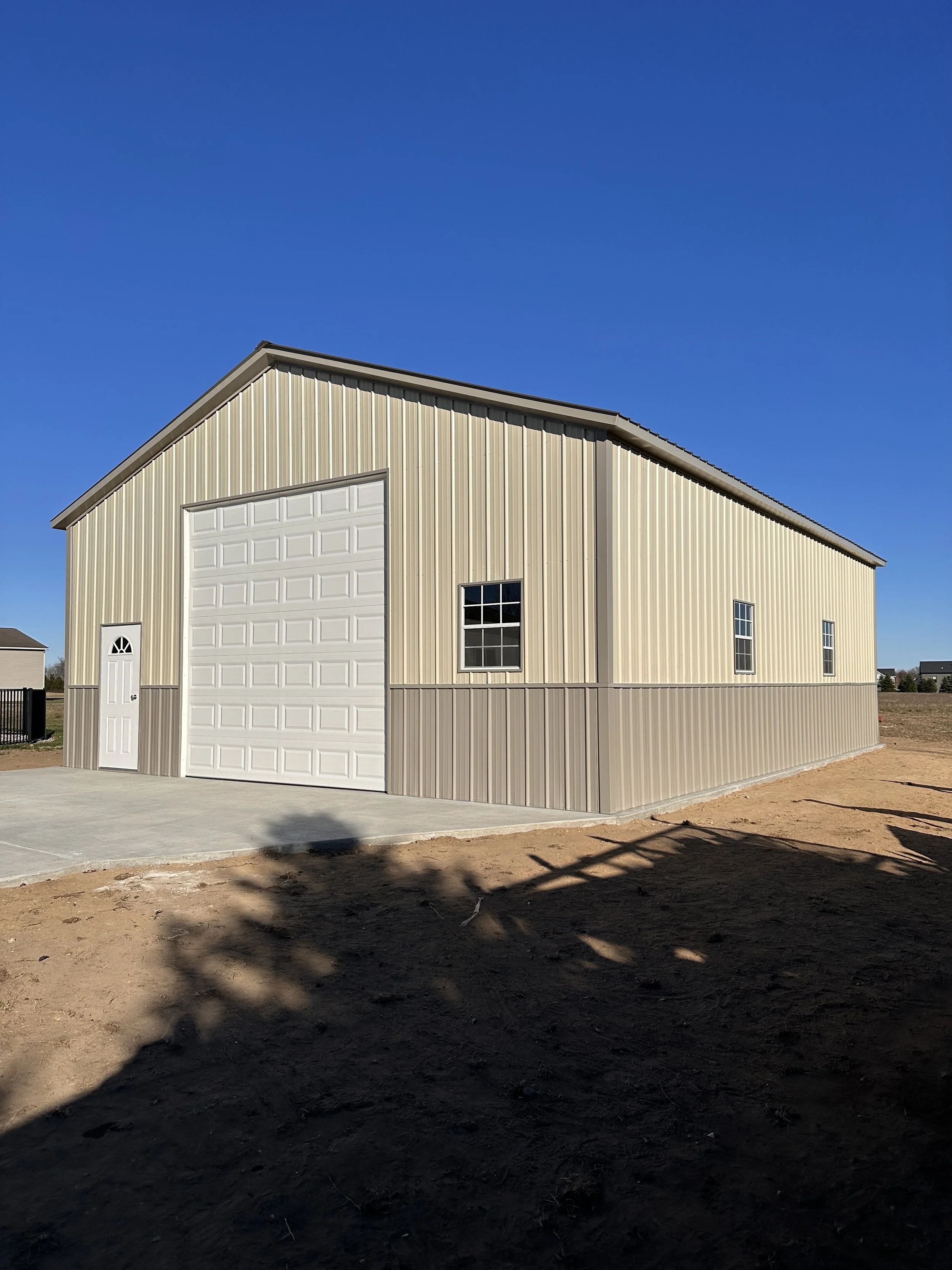 A metal storage building with tan and beige panels, a large white garage door, a side door, and three windows under a clear blue sky.