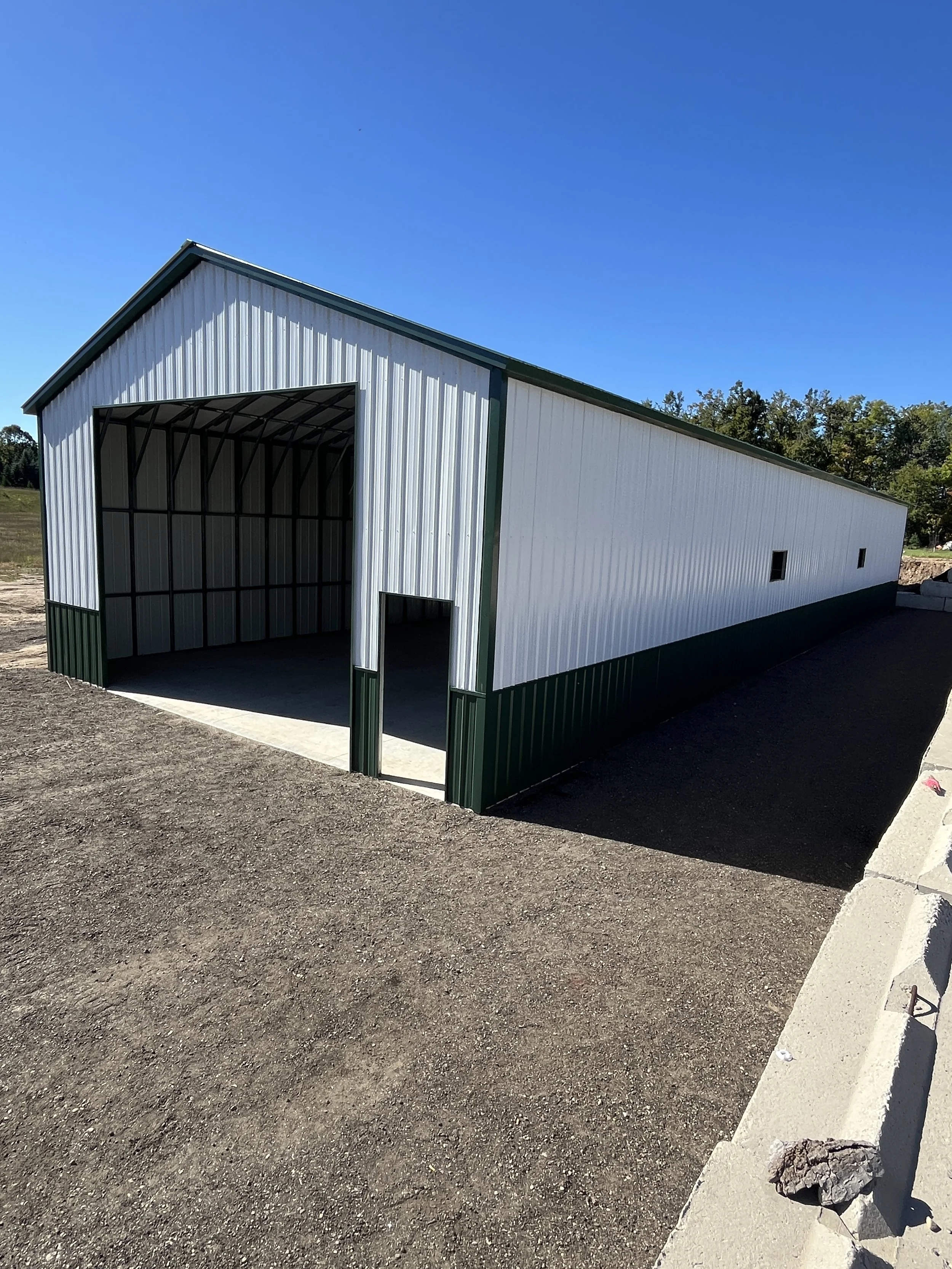 A large metal building, possibly a storage shed or garage, with white walls, green trim, and an open front, situated on a dirt lot under a clear blue sky.
