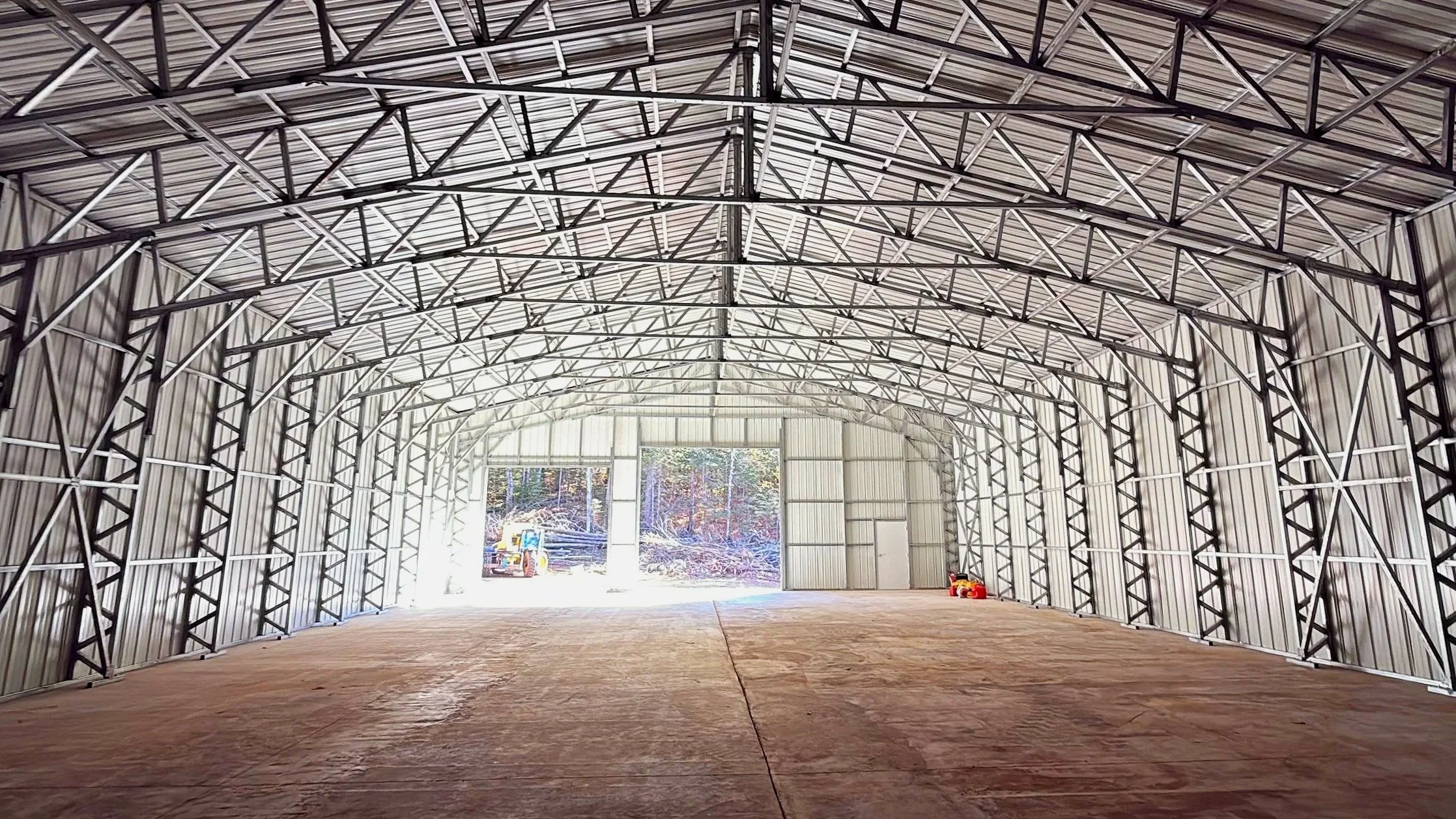 Empty metal building with curved roof and open entrance, wooden floor, construction equipment outside, and trees visible through the opening.