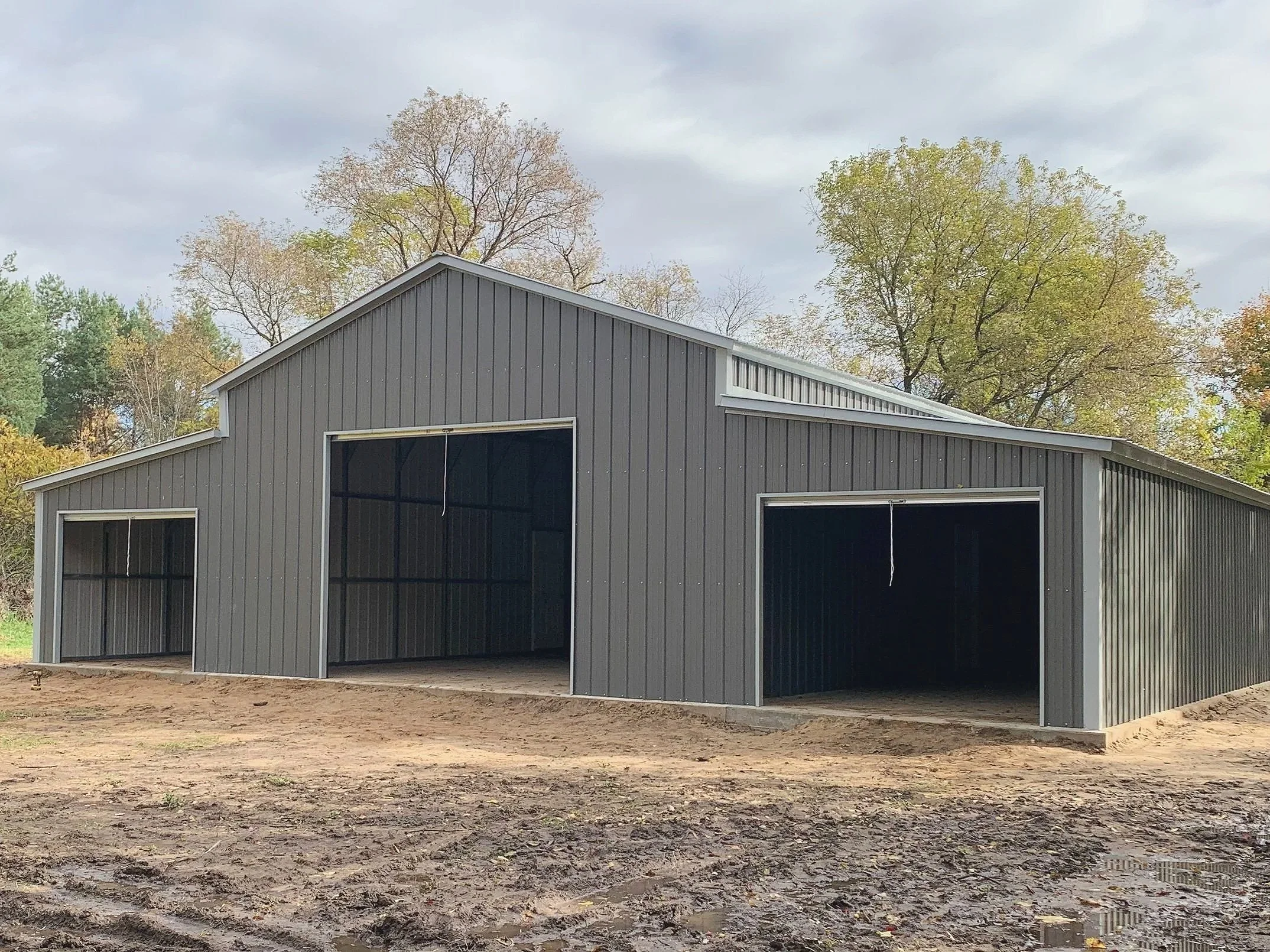 A gray metal barn under construction with three open garage doors, surrounded by dirt and trees in the background.