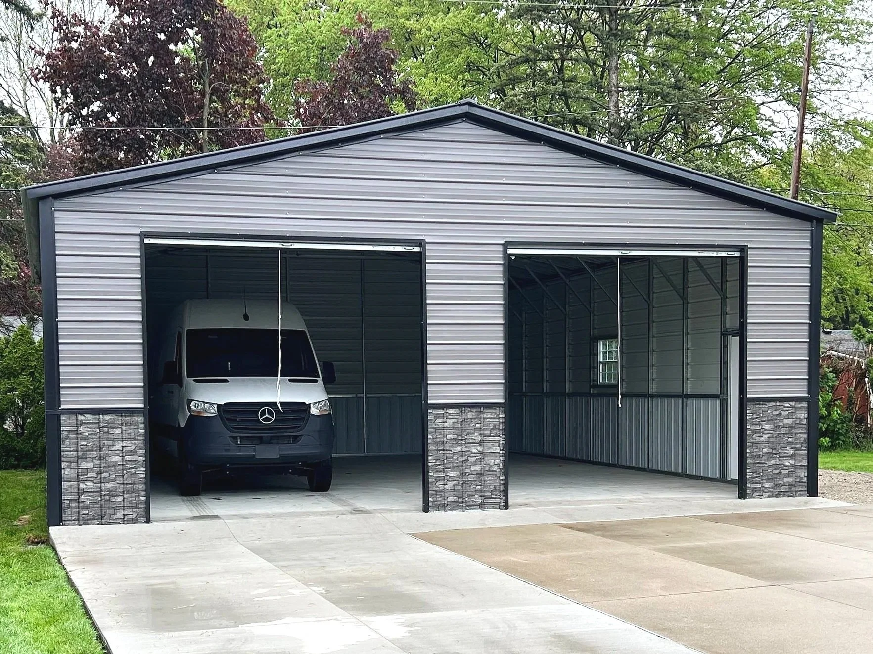 A metal garage with two openings, one of which is open and contains a white Mercedes-Benz van. The garage has gray siding with stone trim at the bottom and is situated on a concrete driveway. There are green trees in the background.