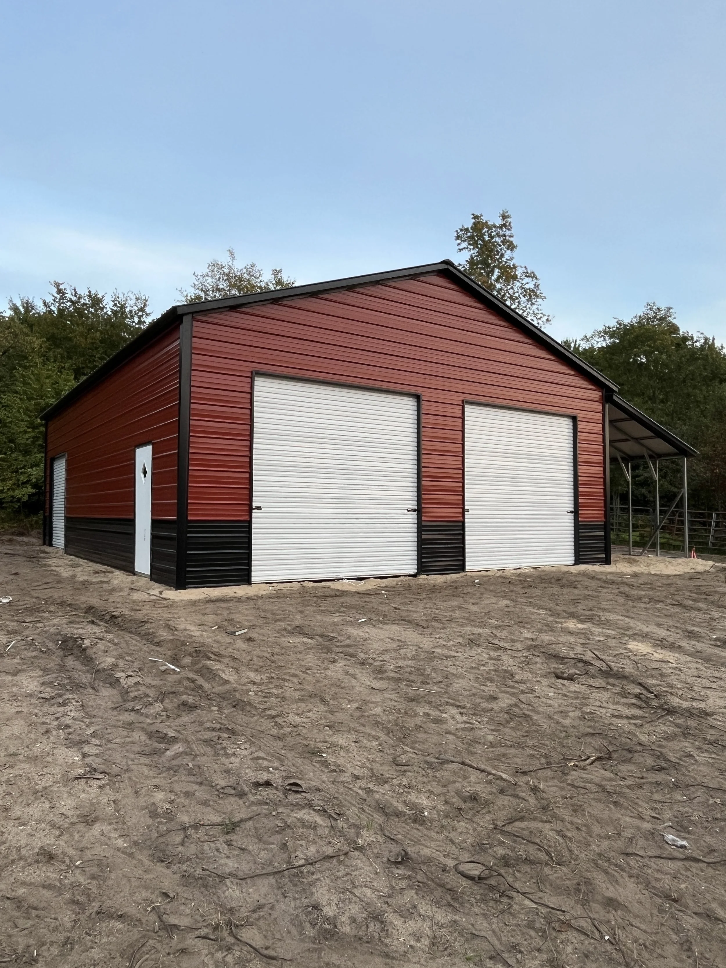 Red and black metal barn with two large white roll-up doors and one smaller white door, located on dirt ground with trees in the background under a clear blue sky.