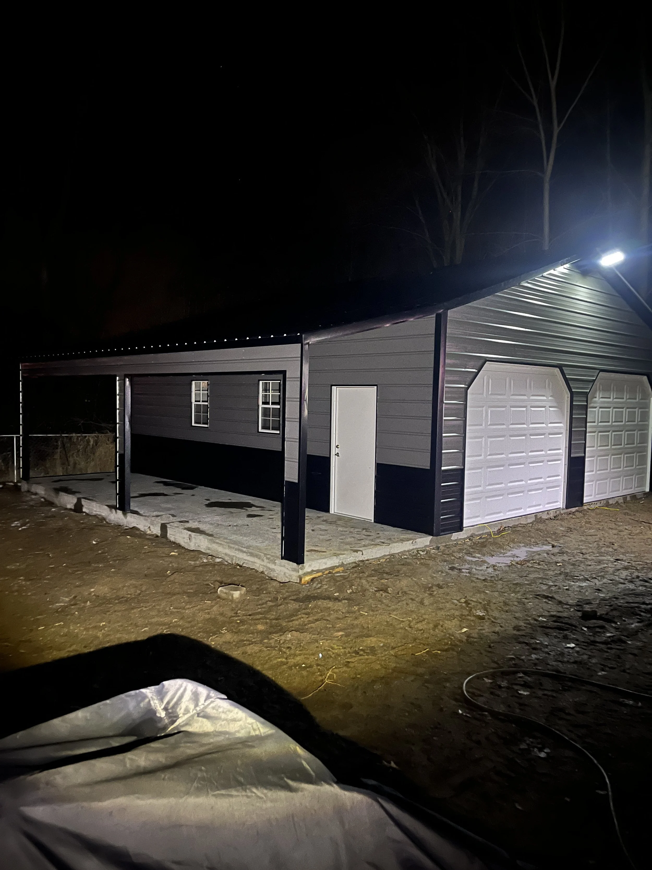 A metal garage with two roll-up doors and one side door, illuminated at night, with a dirt ground in front.
