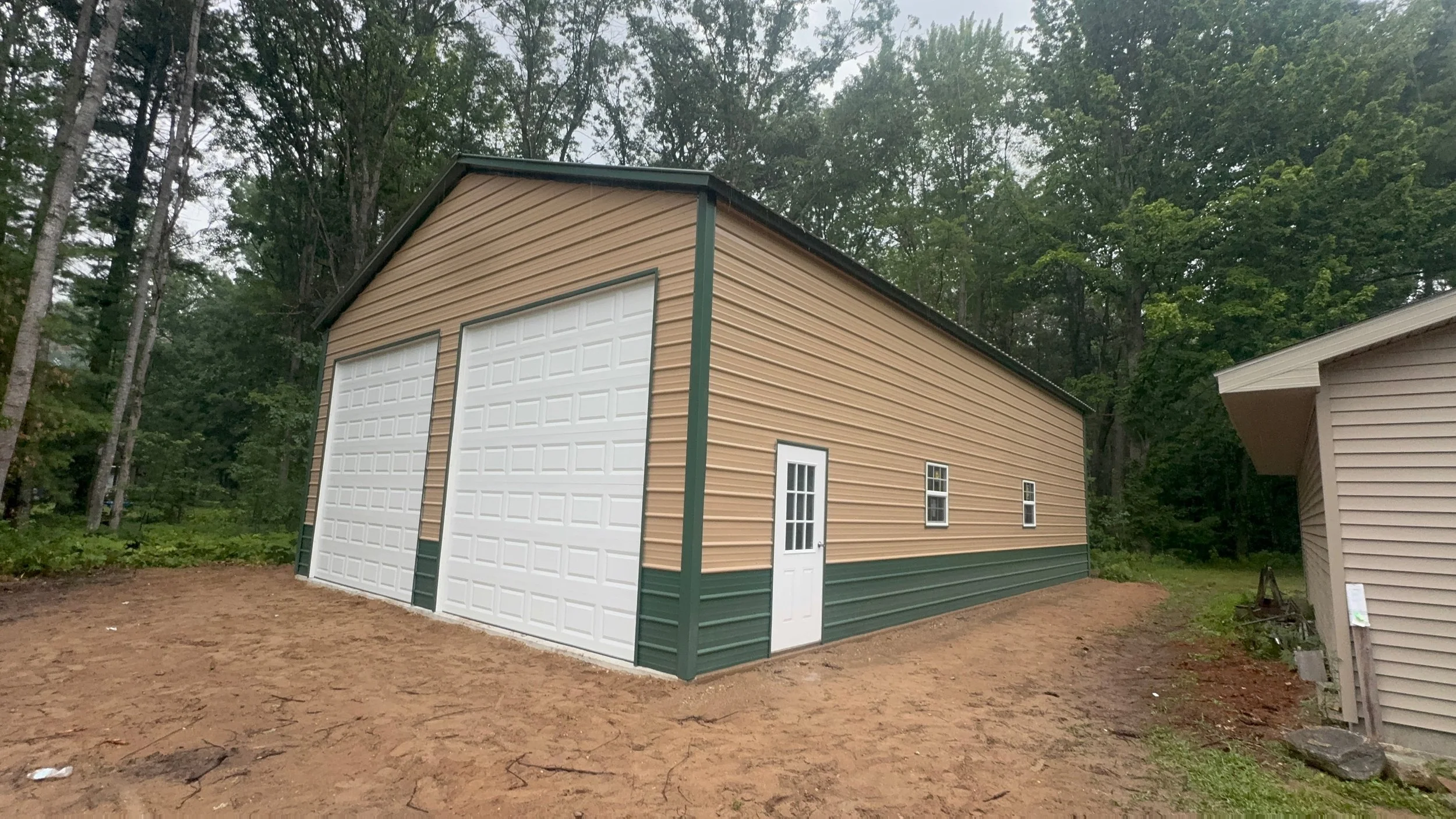 A beige and green metal storage building with two large white garage doors, a small white door, and three small windows, situated in a wooded area with dirt ground.