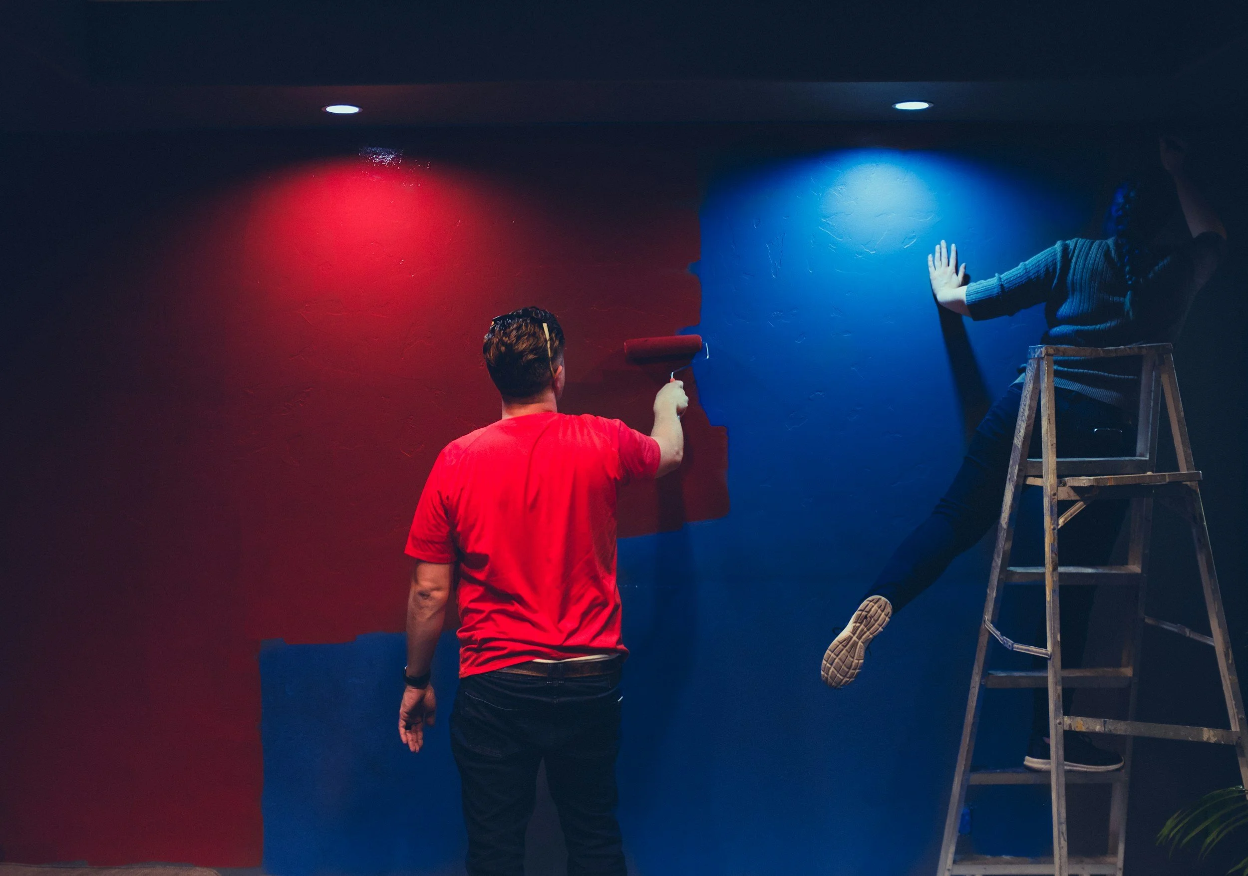 Two people painting a wall with red and blue sections, one on a ladder, using a roller.