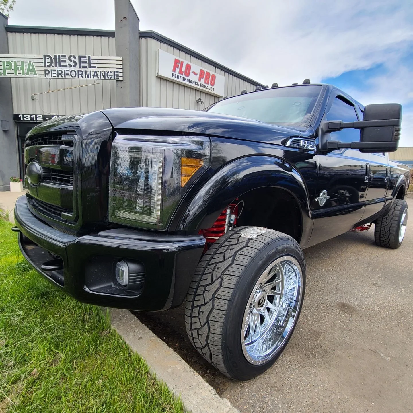 A black lifted Ford F-250 truck with shiny custom wheels parked outside a building with signs for performance exhaust and diesel performance.