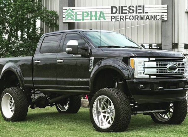 Black lifted Ford F-150 truck with large off-road tires parked on grass in front of a sign that says 'Alpha Diesel Performance'.
