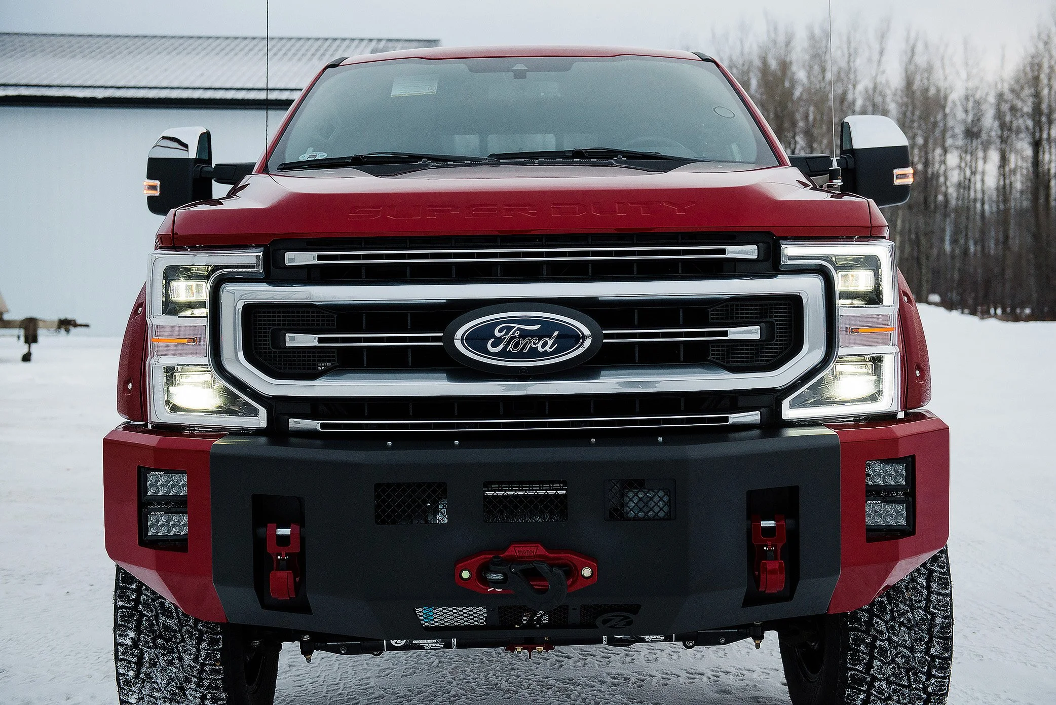 Front view of a red Ford Super Duty truck with a black grille and LED headlights, parked on snow with trees in the background.