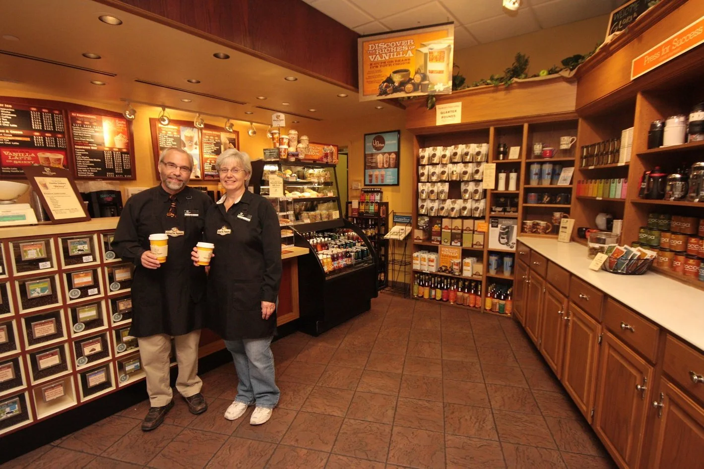 Scott and his wife Shirley smiling while holding cups of coffee. Behind them is their coffee shop, "Gloria Jeans" that was located in the Oakwood Mall in Eau Claire.