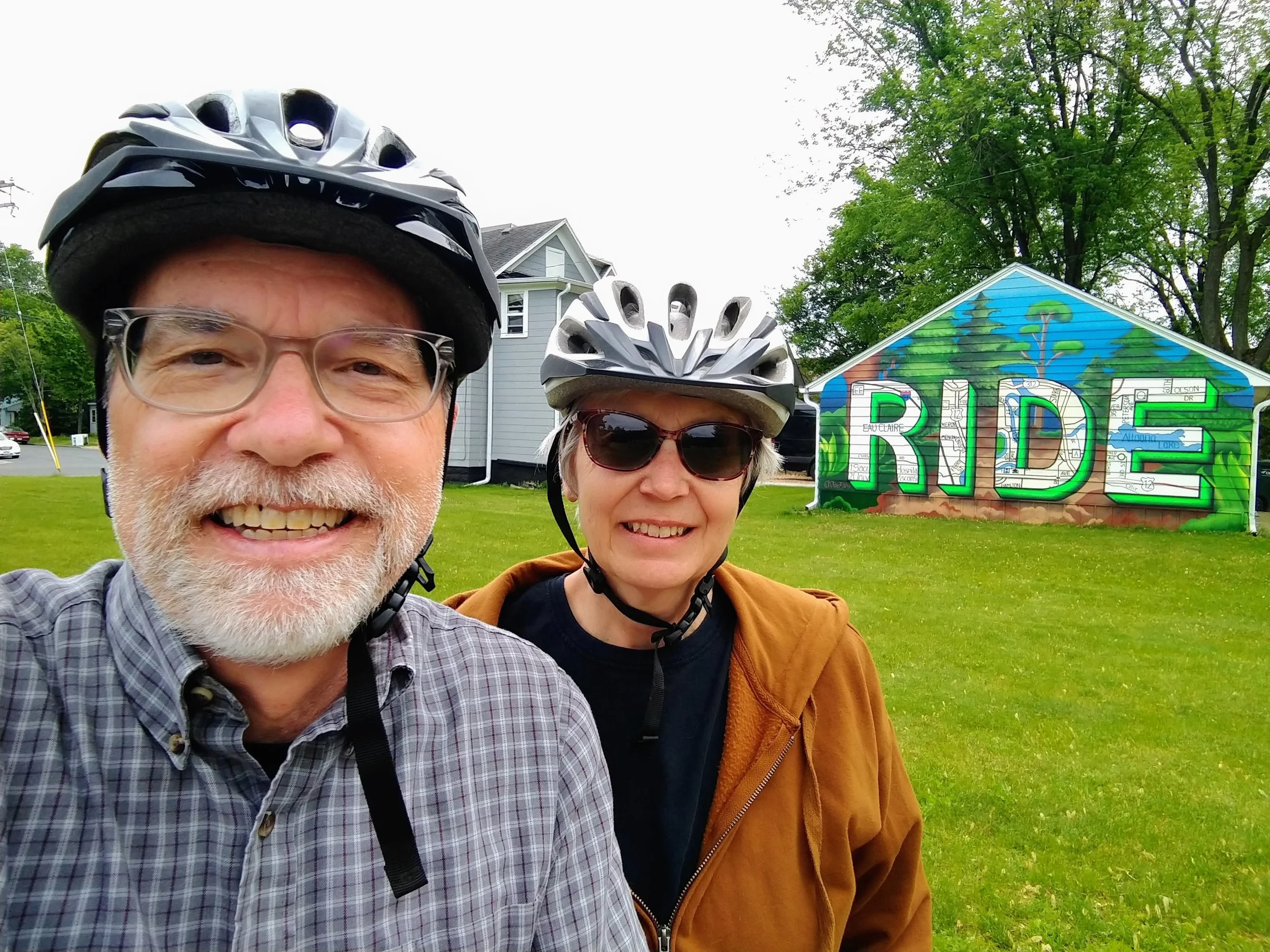 Scott and Shirley smiling at the camera. They are wearing bike helmets and are in front of a garage with a mural that says, "Ride".