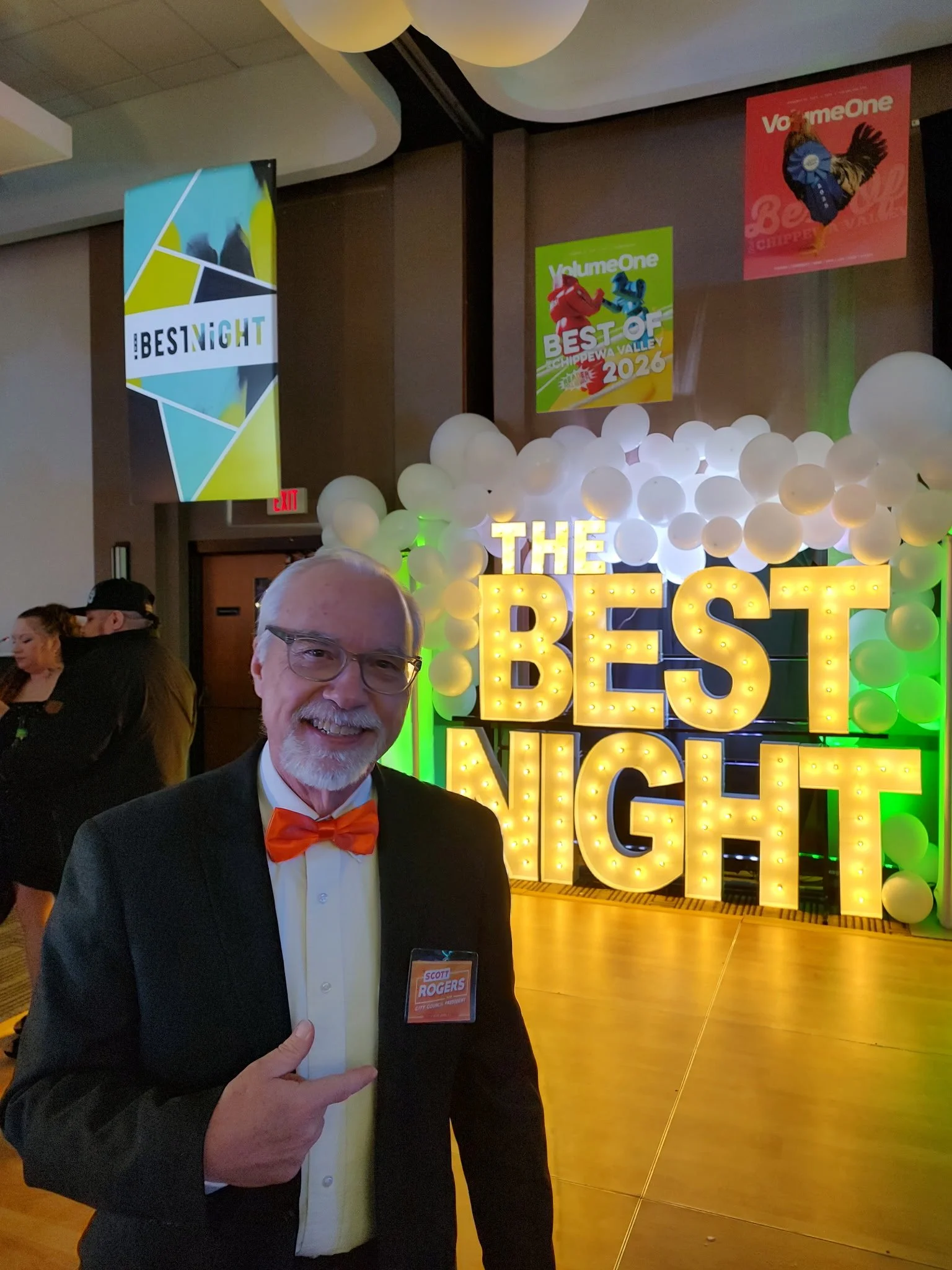 Scott wearing an orange bow tie in front of a light up sign that says, "The Best Night"