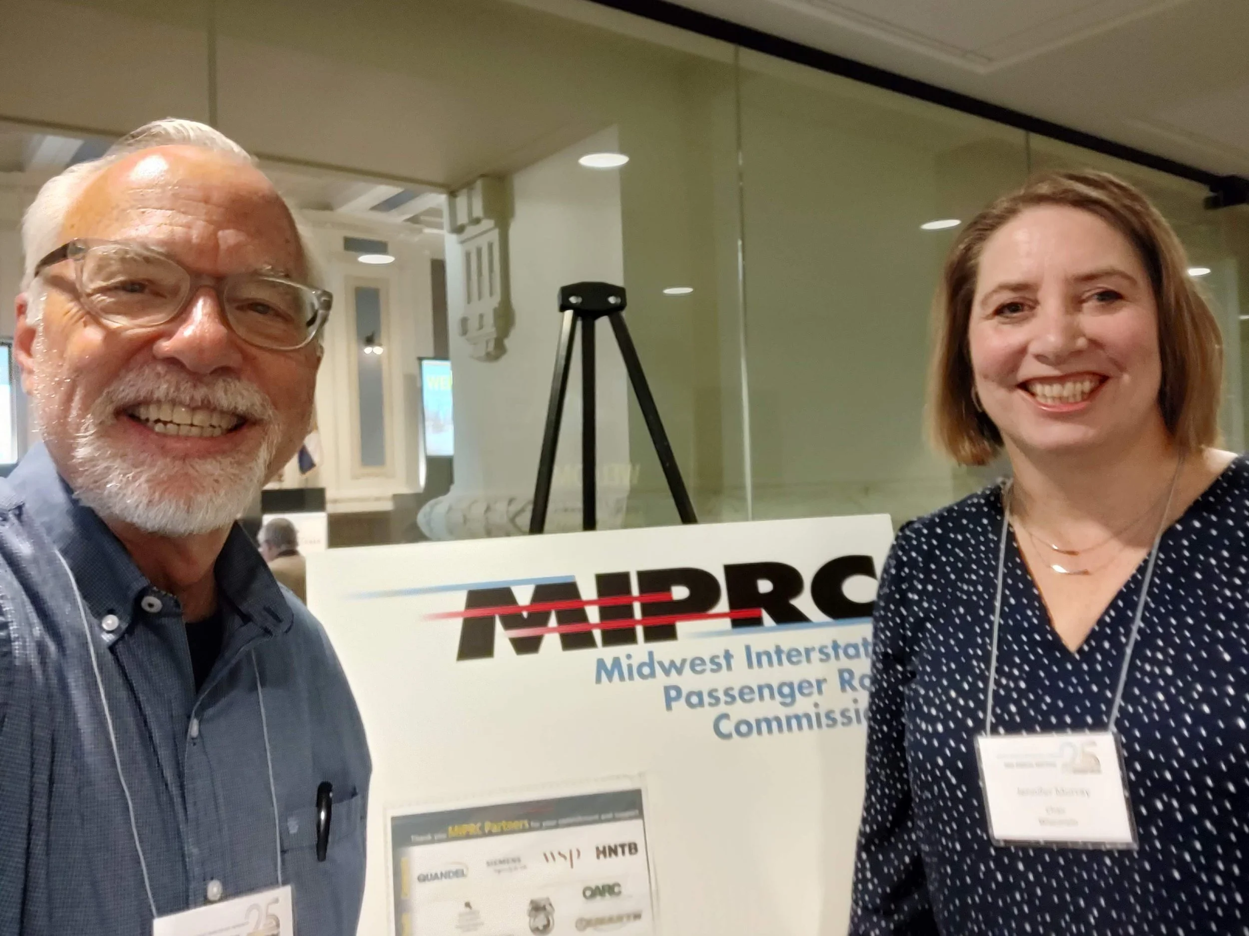Scott and Jennifer Murray smile at the camera. Behind them is a poster board that says, Midwest Interstate Passenger Rail Commission.