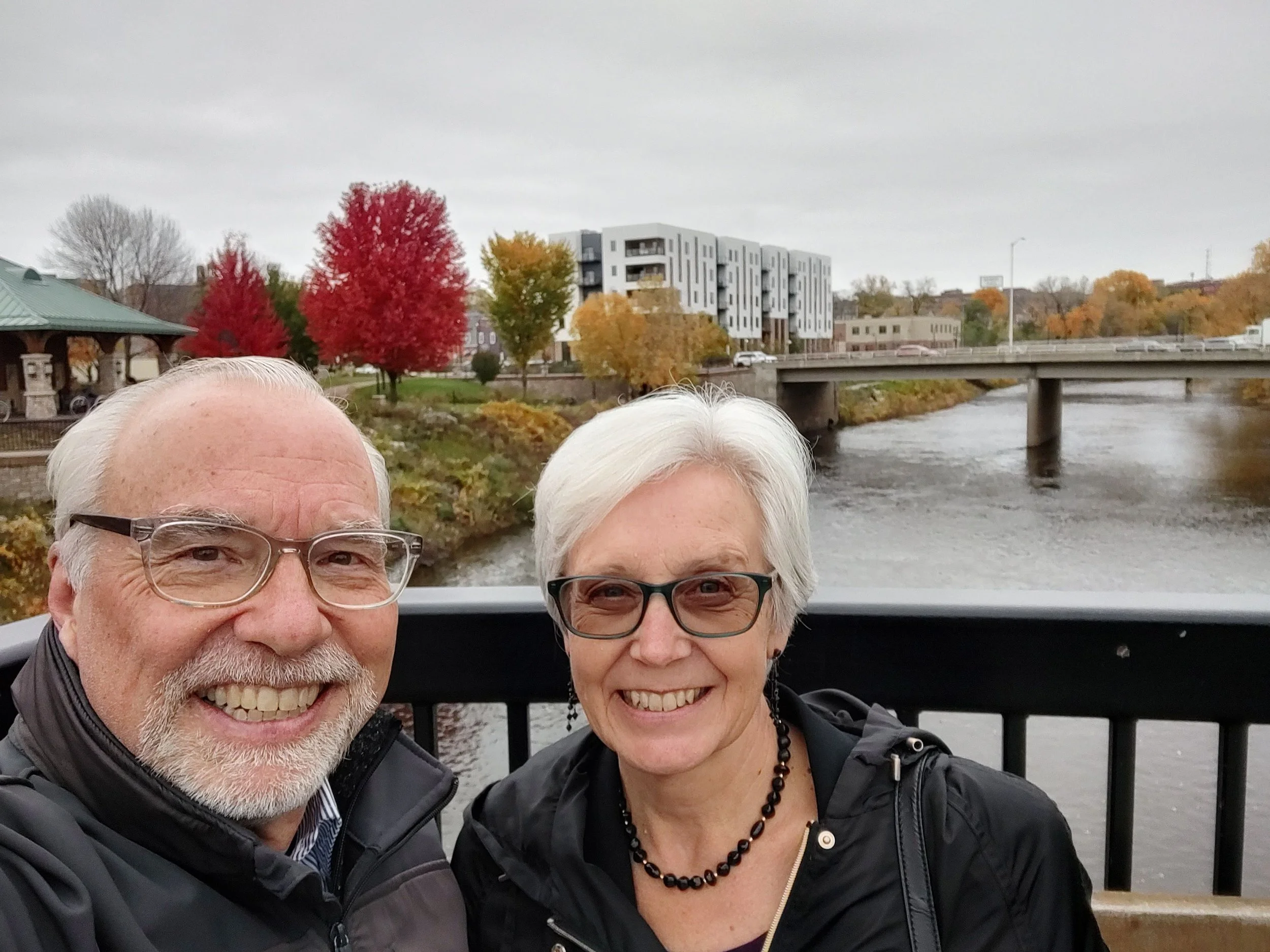 Scott and his wife Shirley smiling at the camera. Behind them is the a white apartment building, river and bridges in downtown Eau Claire where they live.