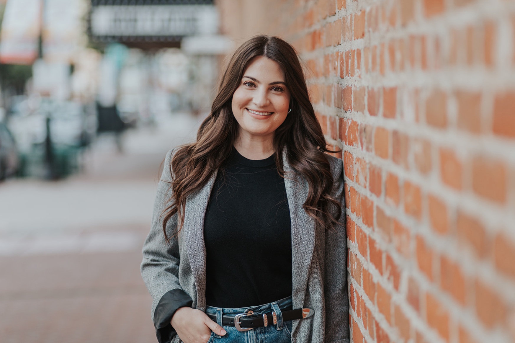 A woman with long wavy brown hair smiling and leaning against a red brick wall on an urban street.
