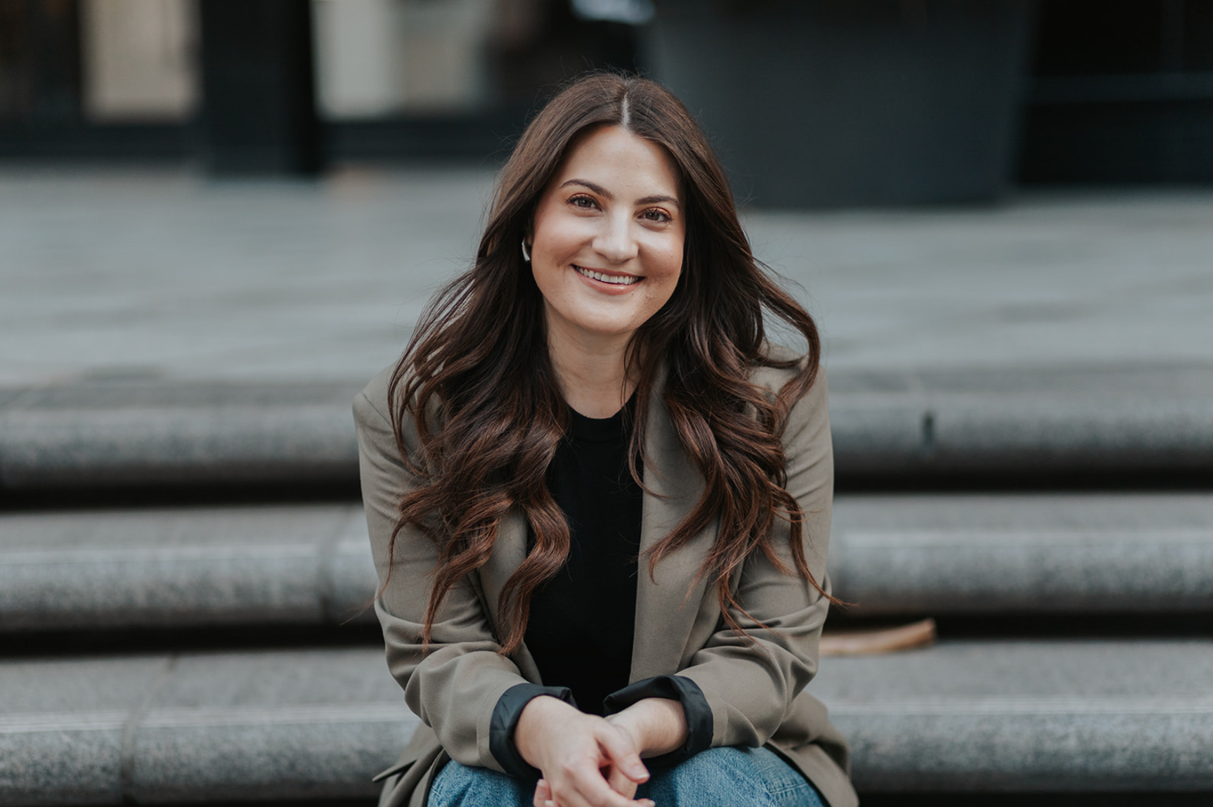 A woman with long brown wavy hair sitting on outdoor steps, smiling, wearing a green blazer over a black top.