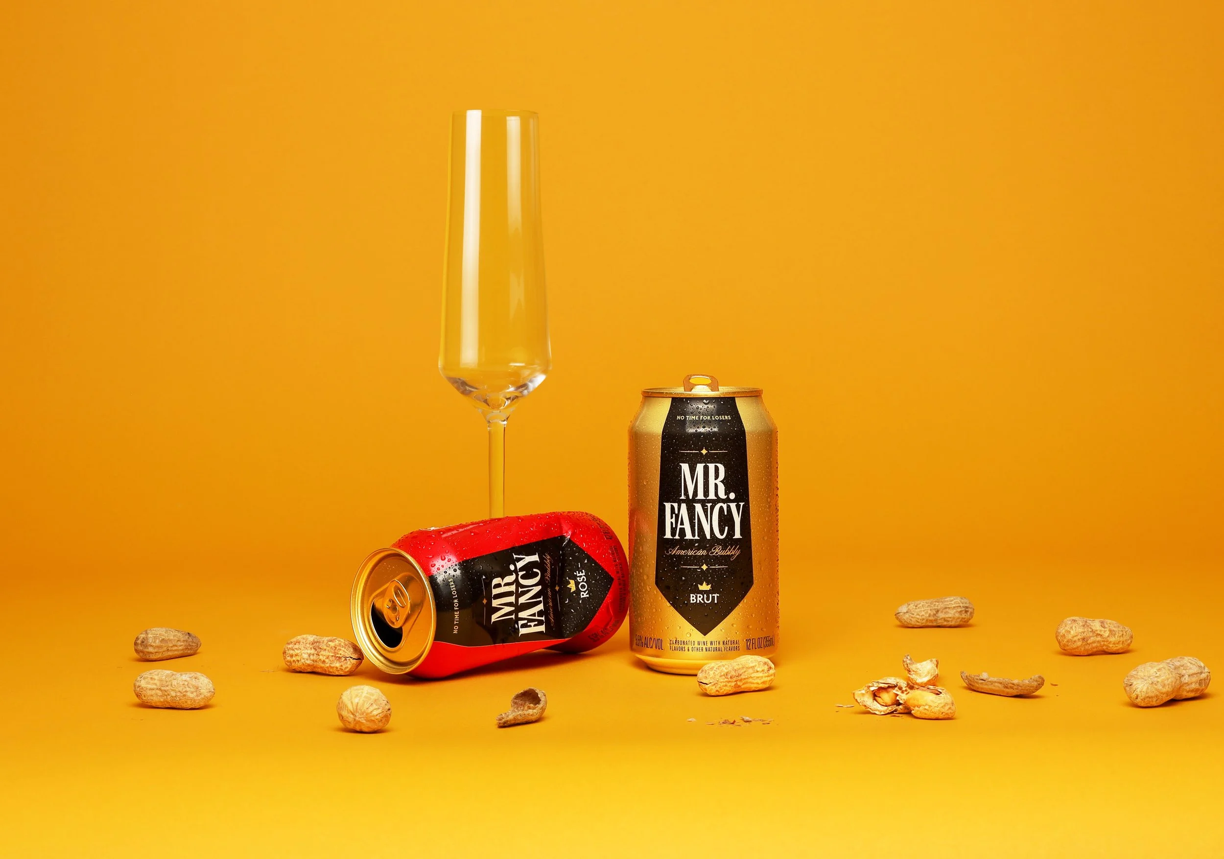 A tall champagne flute glass and two cans of Mr. Fancy sparkling wine on a yellow background, with scattered peanuts and peanut shells around them.