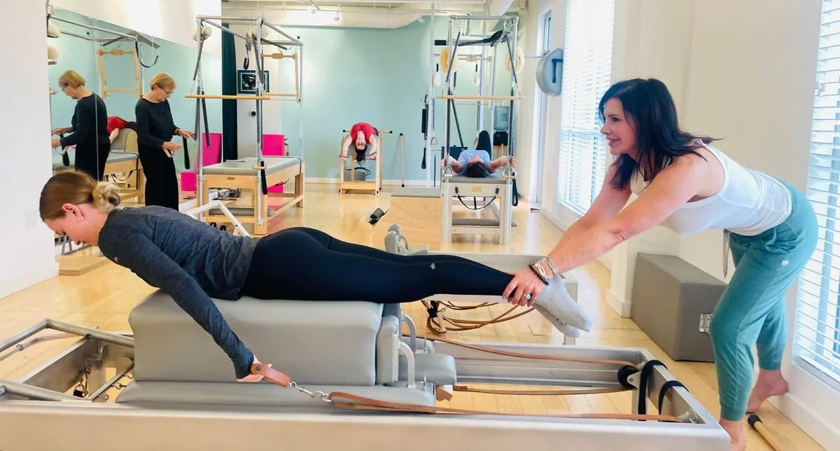 A woman is assisting another woman with a Pilates reformer machine in a fitness studio. The woman on the reformer is lying face down, while the instructor holds her legs. In the background, people are doing exercises on various equipment.