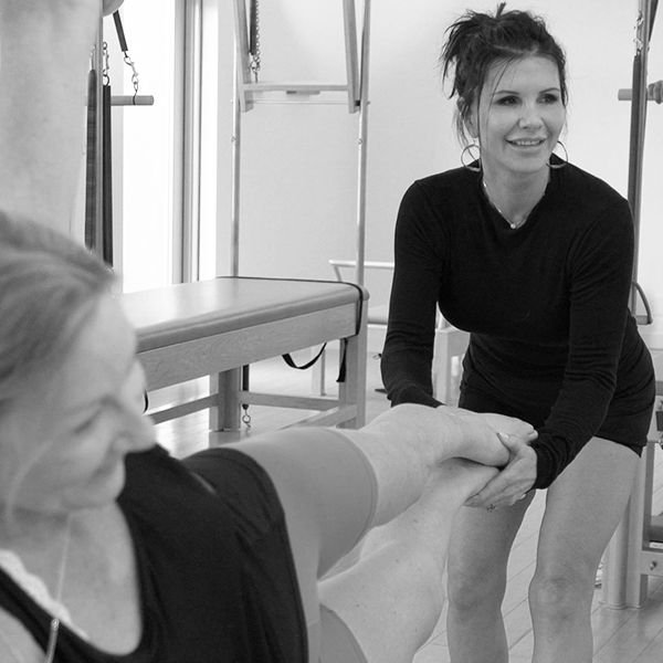 A woman in a black shirt sitting on a chair beside a patient in a healthcare setting, holding the patient's hand and smiling.