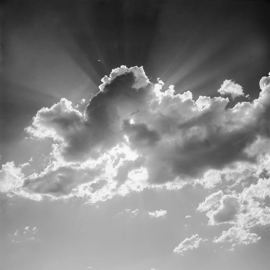 Black and white photo of a cloudy sky with the sun behind the clouds, creating sun rays.