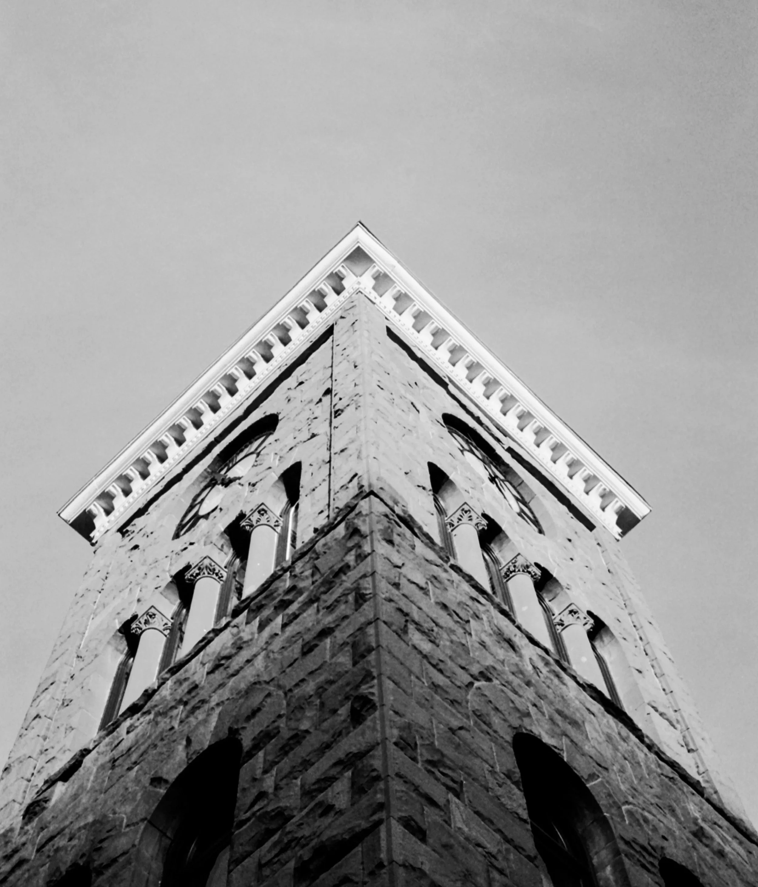 Black and white photo of a historic stone building corner with arched windows and decorative architectural details at the top, viewed from below, against a plain sky.