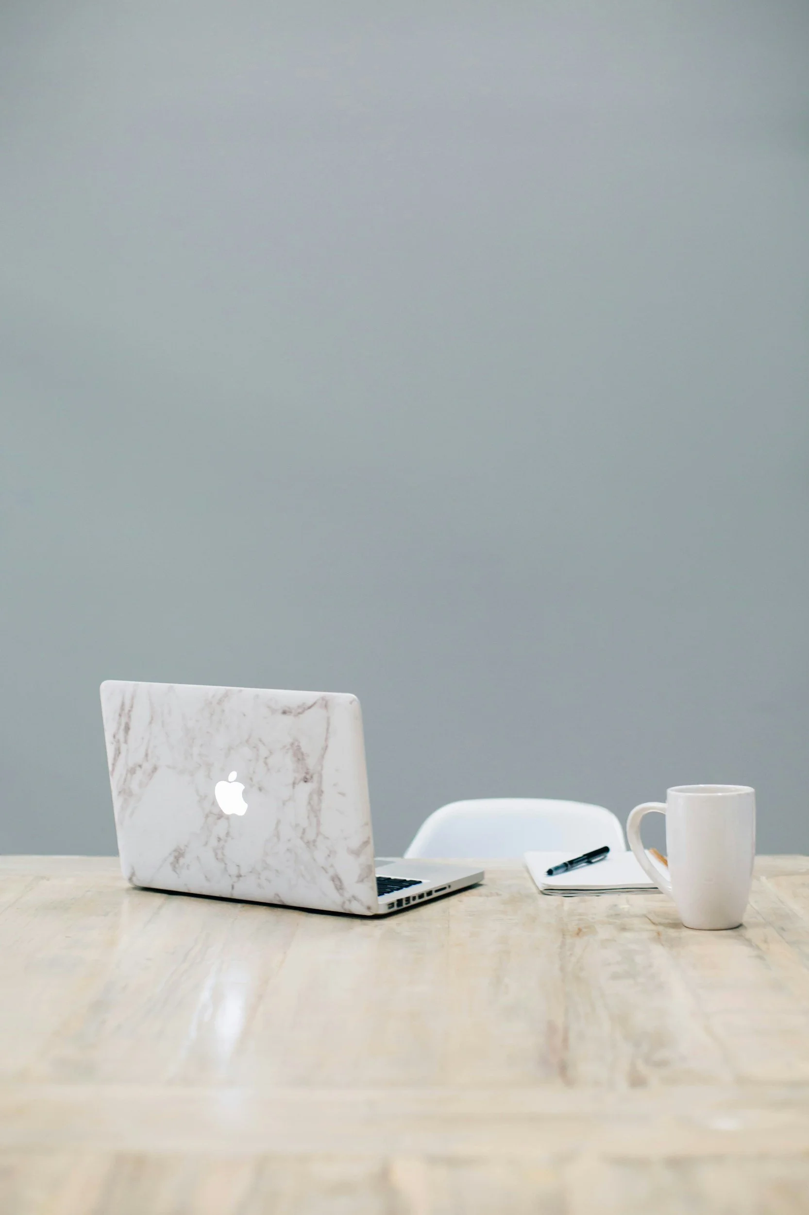 Office desk with a marble-patterned MacBook, a white chair, a notepad, a black pen, and a white mug on a light wood table against a plain gray wall.