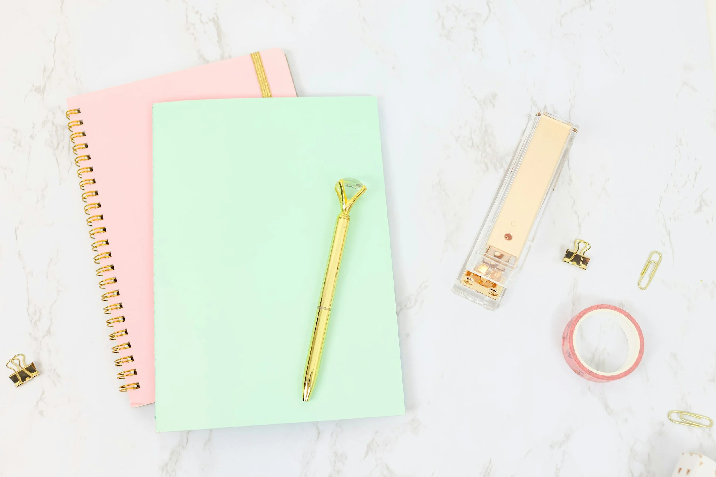 Flat lay of office supplies including pastel-colored notebooks, a gold pen with a gemstone top, a correction tape, a screw puncher, paper clips, binder clips, and decorative tape on a white marble surface.