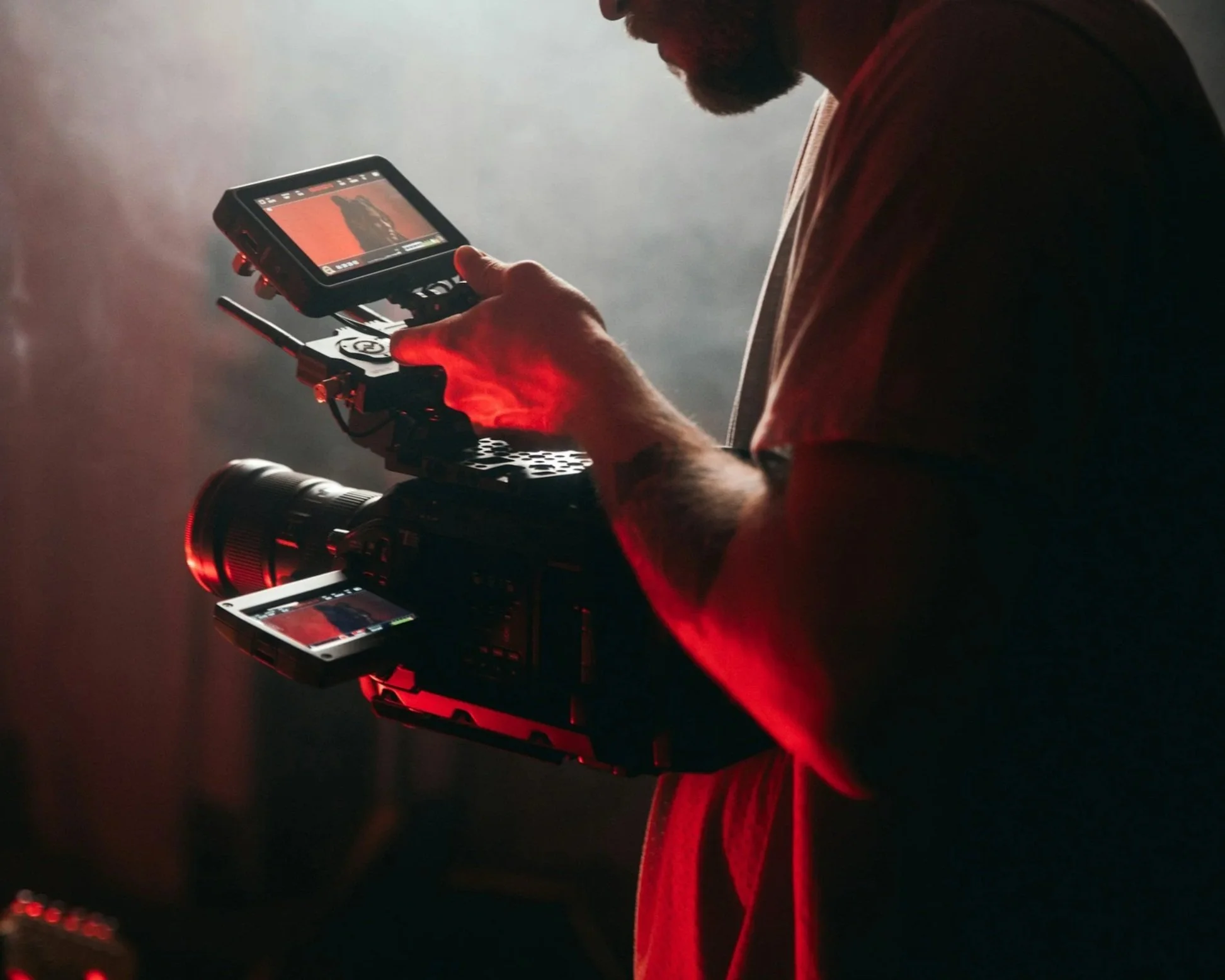 A person operating a professional video camera with dual screens, one on top and one on the side, in a dimly lit environment with red lighting.