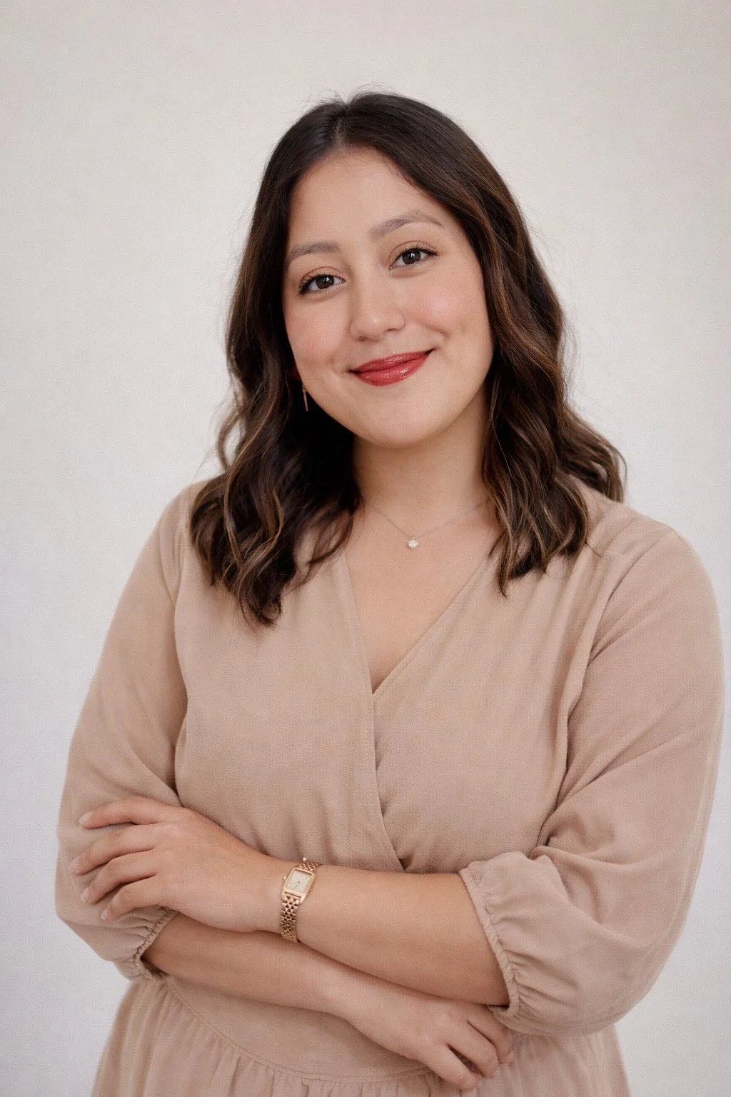 A woman with shoulder-length wavy brown hair, wearing a beige dress, gold watch, delicate necklace, and earring, smiling with arms crossed against a plain white background.