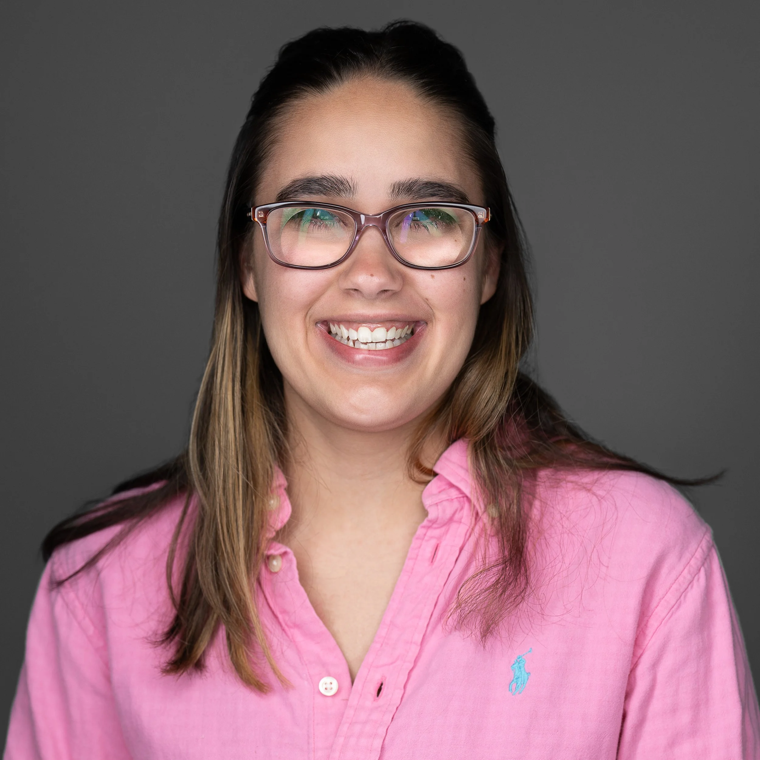 A young woman with glasses smiling, wearing a pink collared shirt against a gray background.