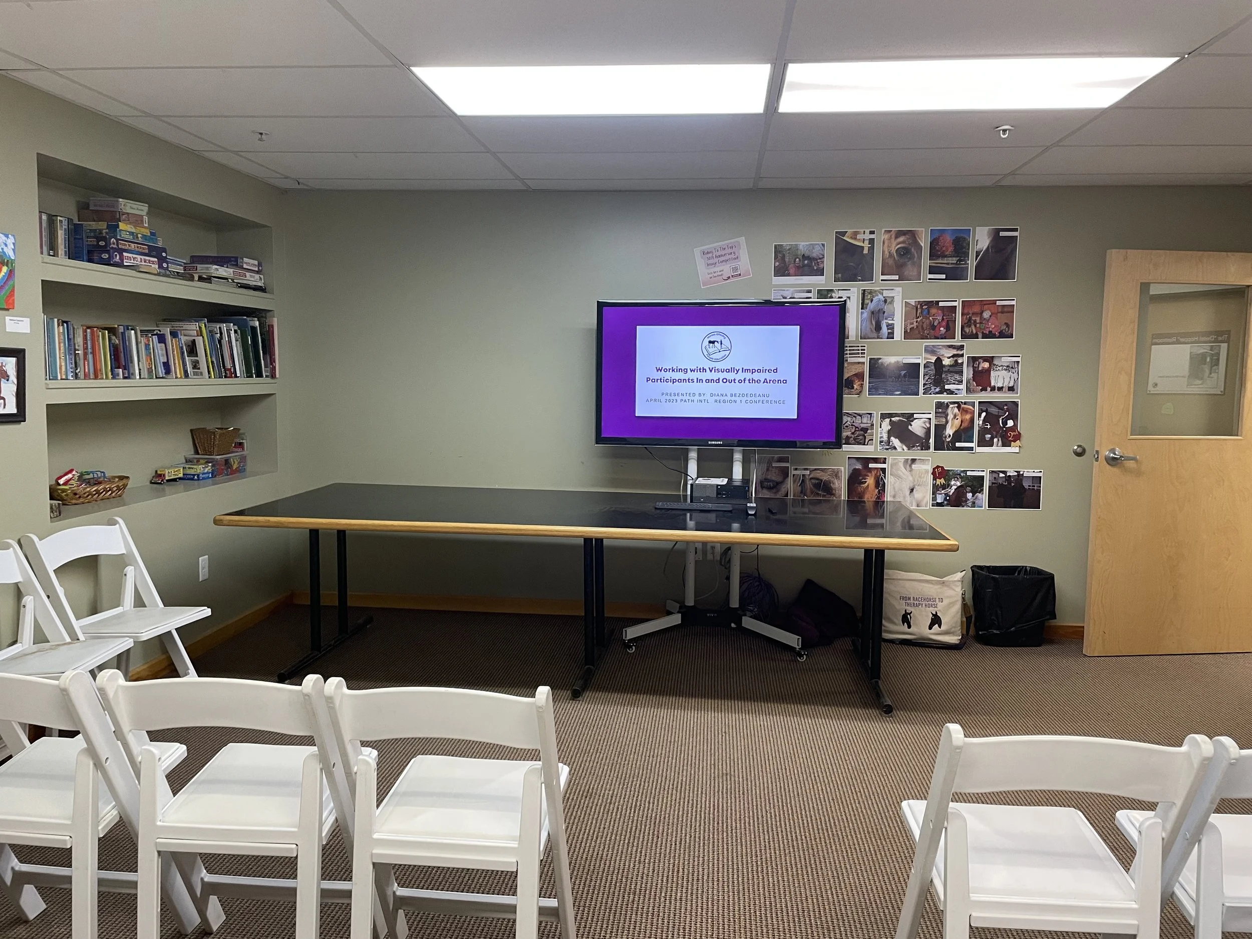 A classroom or conference room with chairs arranged facing a large TV screen on a table. The screen displays a presentation titled "Working with Visually Impaired Participants in and Out of the Arena." There are bookshelves, photos on the wall, and a wooden door to the right.
