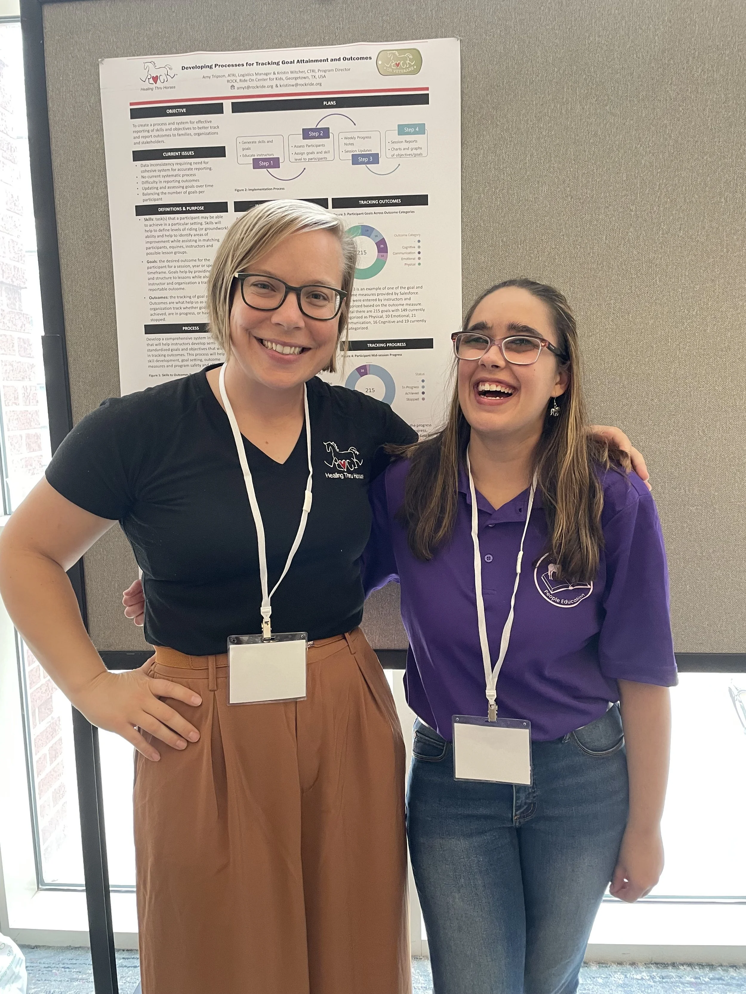 Two women smiling and posing for a photo in front of a research poster; the woman on the left has short blonde hair, glasses, a black T-shirt, and brown pants. The woman on the right has long brown hair, glasses, and is wearing a purple polo shirt and jeans. Both are wearing conference name tags.