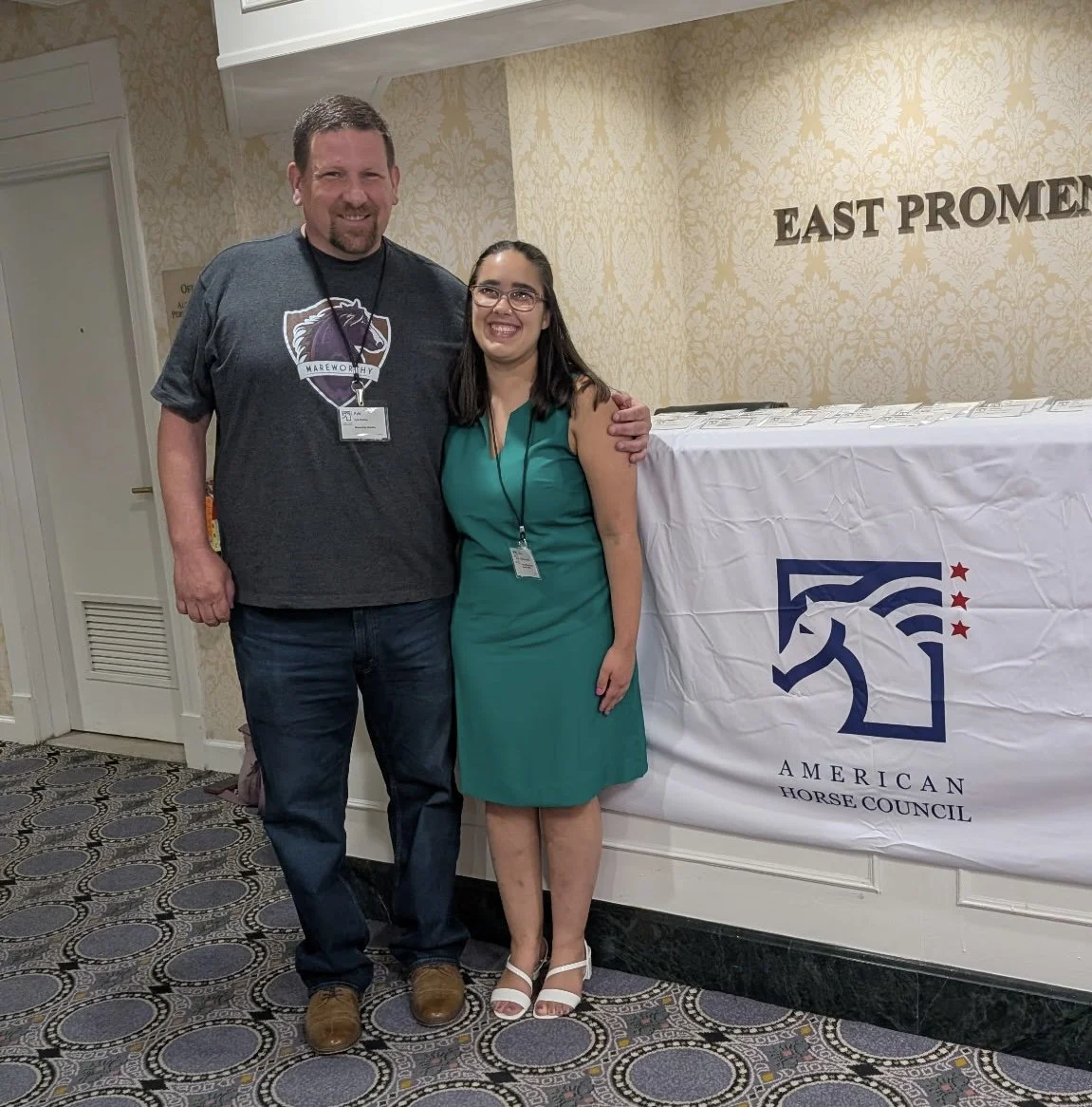 Two people standing together at a conference, smiling, with a banner displaying the American Horse Council logo in the background.