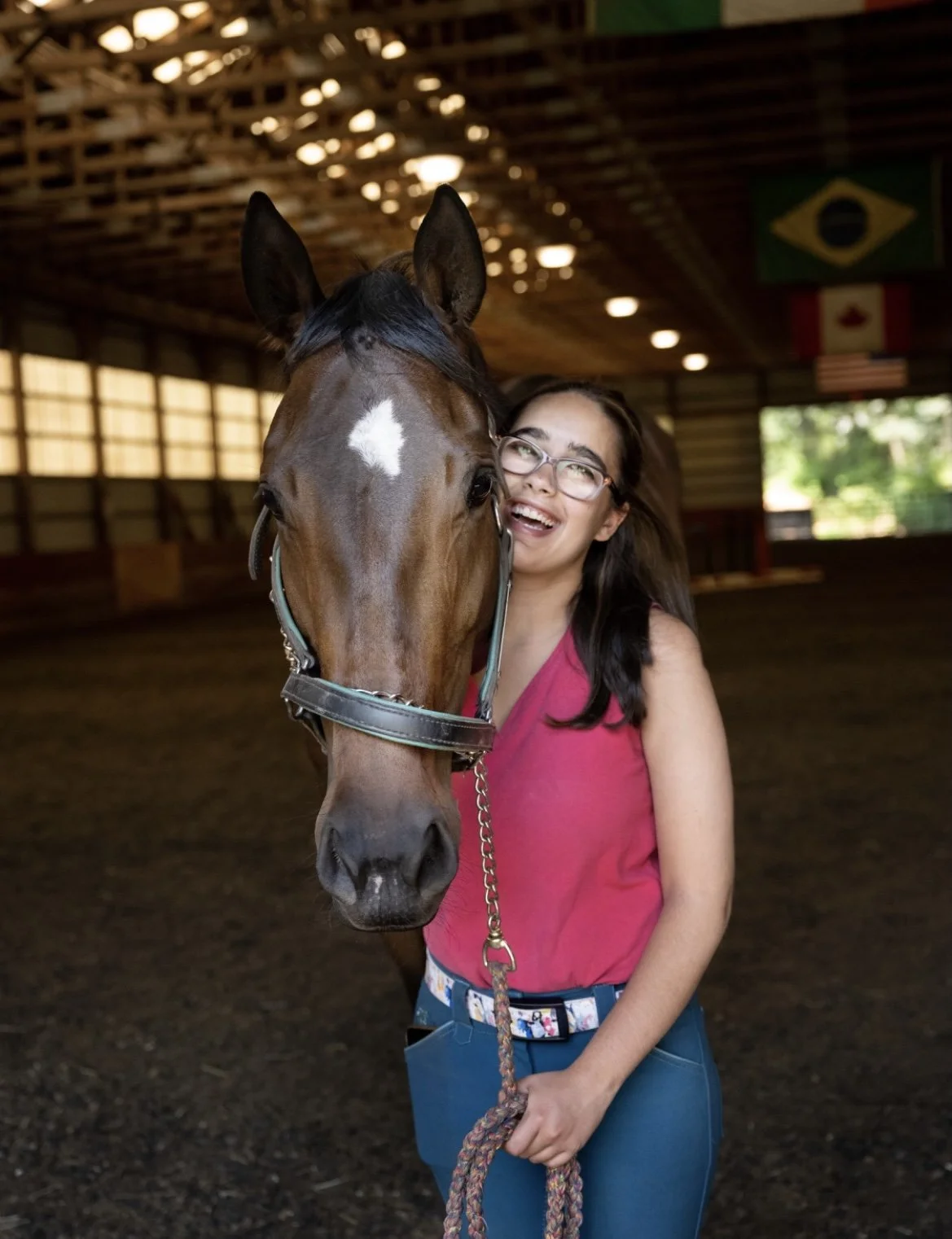 A woman with glasses and long dark hair wearing a pink sleeveless top and blue pants holding a brown horse with a white star on its forehead inside a covered riding arena.