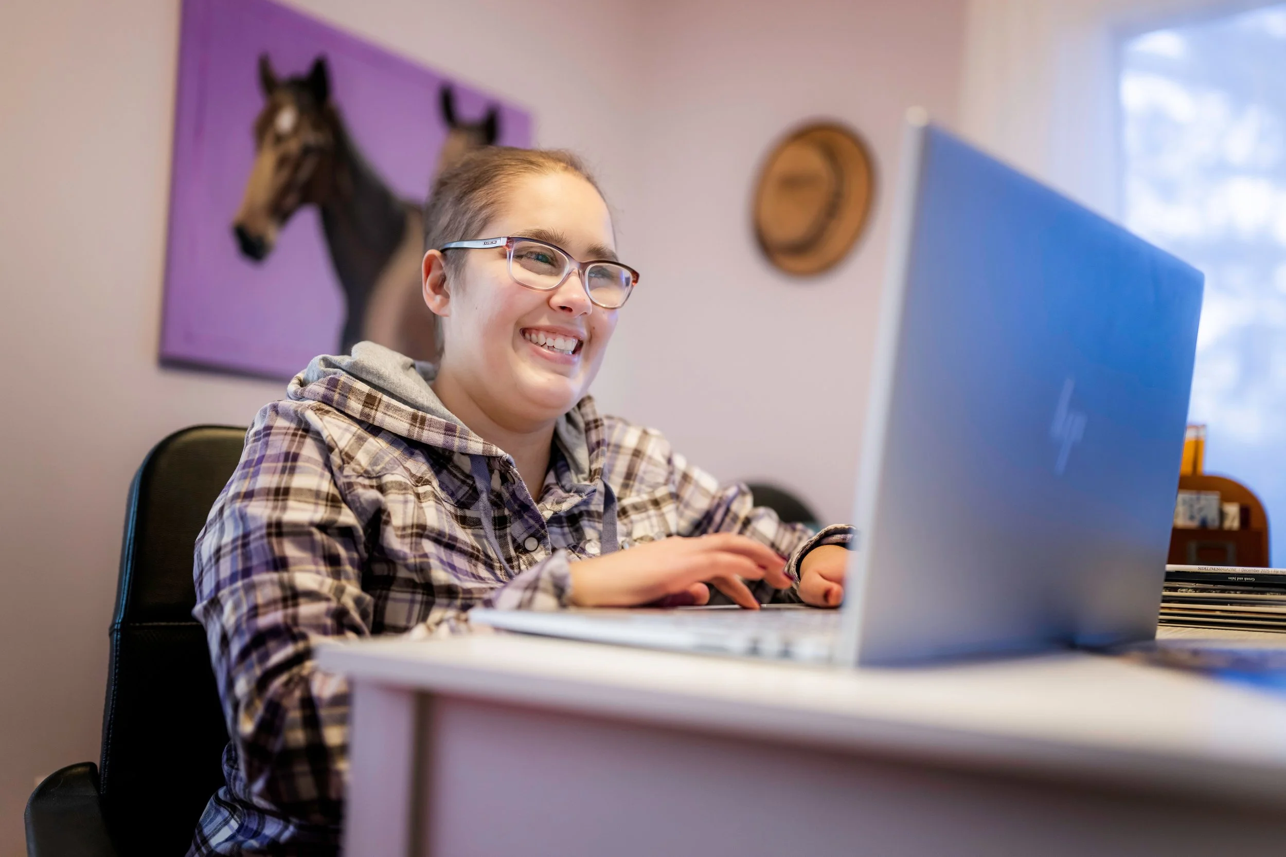 A young woman with glasses and a plaid hoodie sitting at a desk, smiling at her laptop.