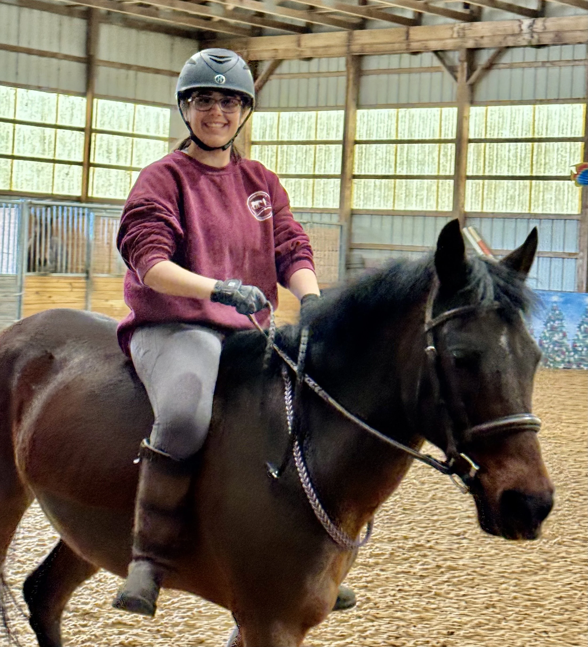 A woman riding a brown horse inside an indoor riding arena, wearing a helmet, glasses, a maroon sweatshirt, and light-colored riding pants.