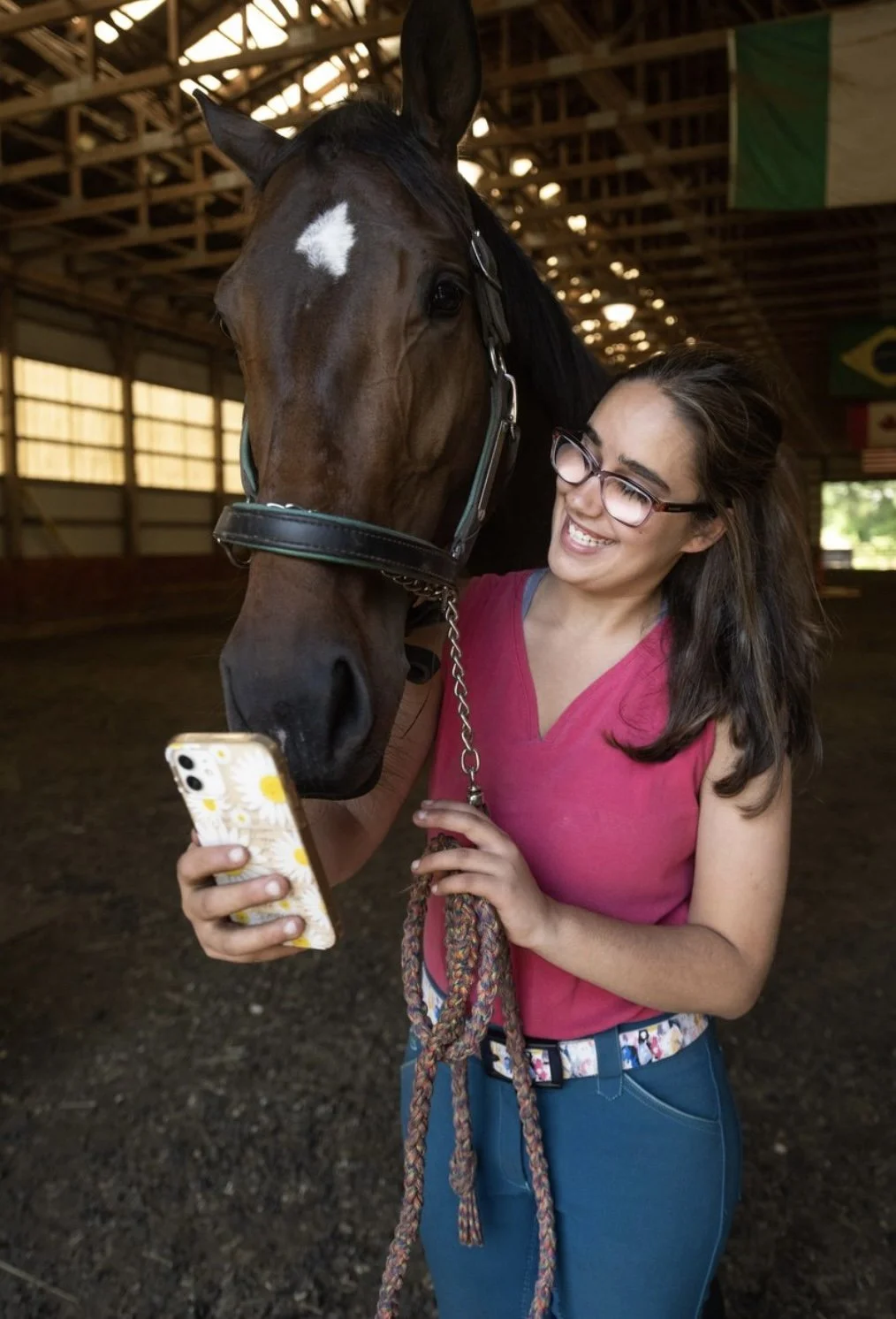 A woman with glasses in a pink top and blue jeans is smiling and taking a selfie with her horse inside a barn.