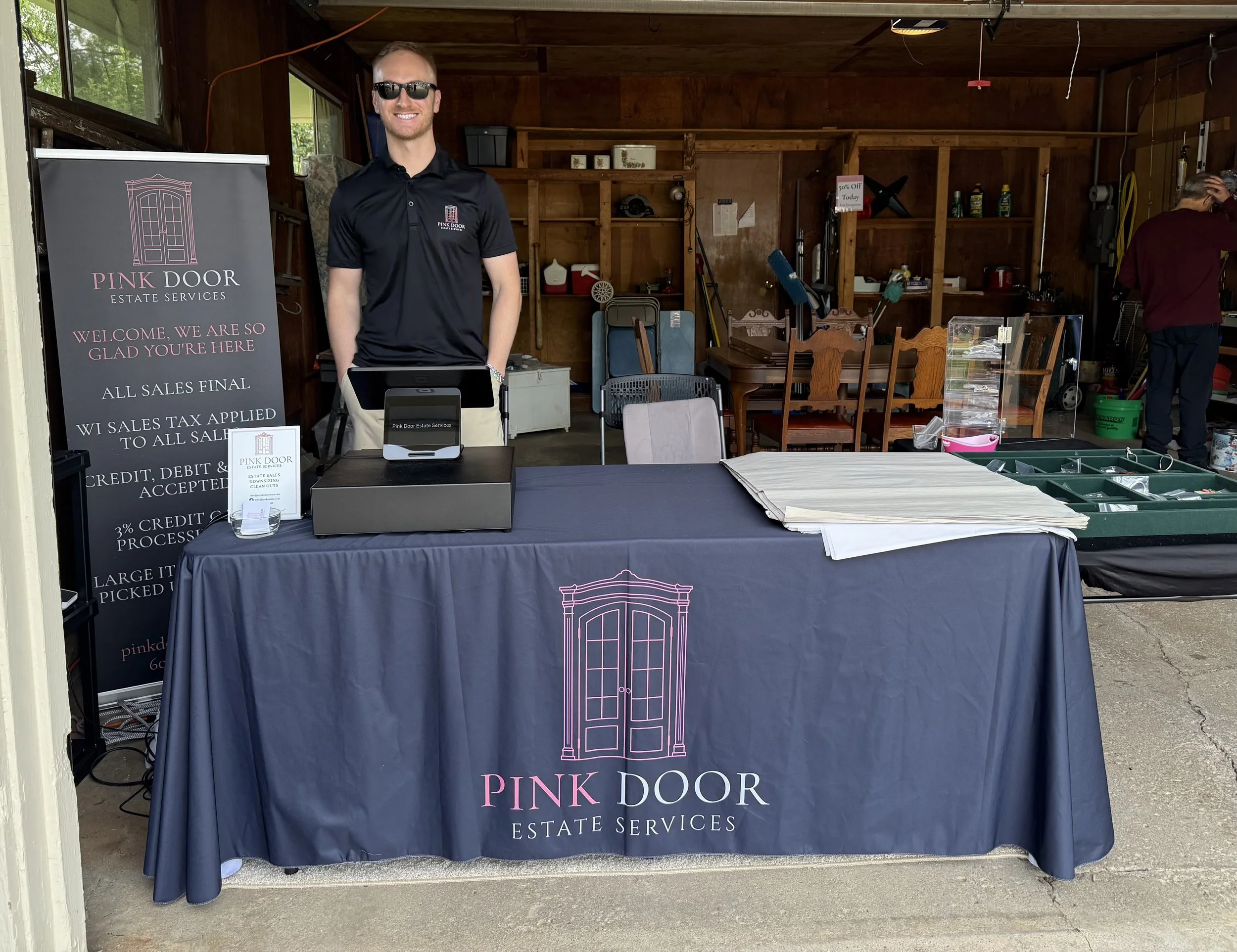 A man wearing sunglasses and a black polo shirt standing behind a table with a pink door estate services sign, inside a wooden shelter. There are chairs, tools, and another person in the background.