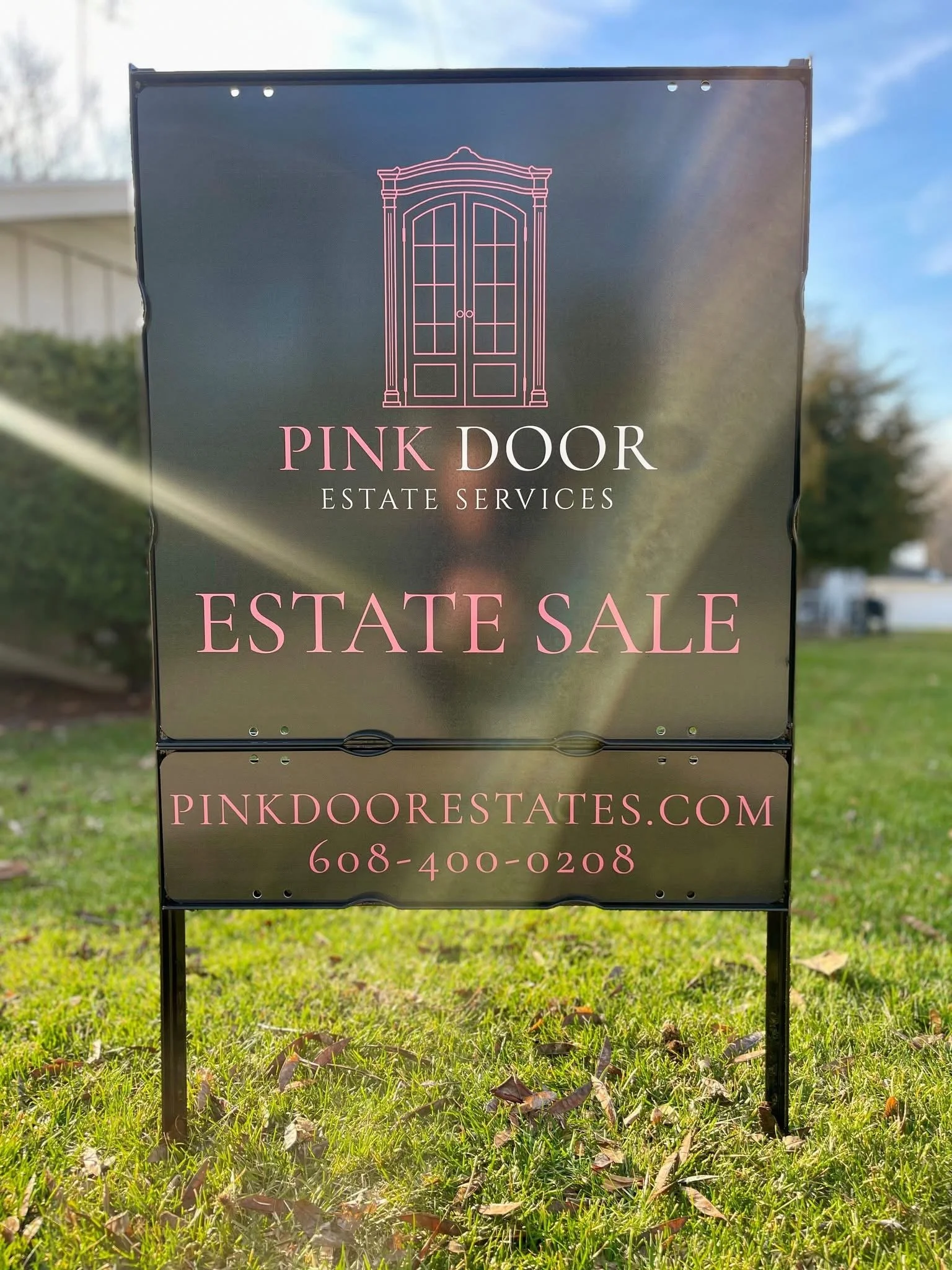 Sign advertising estate sale by Pink Door Estate Services, featuring a pink outline of a cabinet or double door at the top, with the text 'Pink Door Estate Services,' 'Estate Sale,' the website pinkdoorestates.com, and a phone number 608-400-0208.