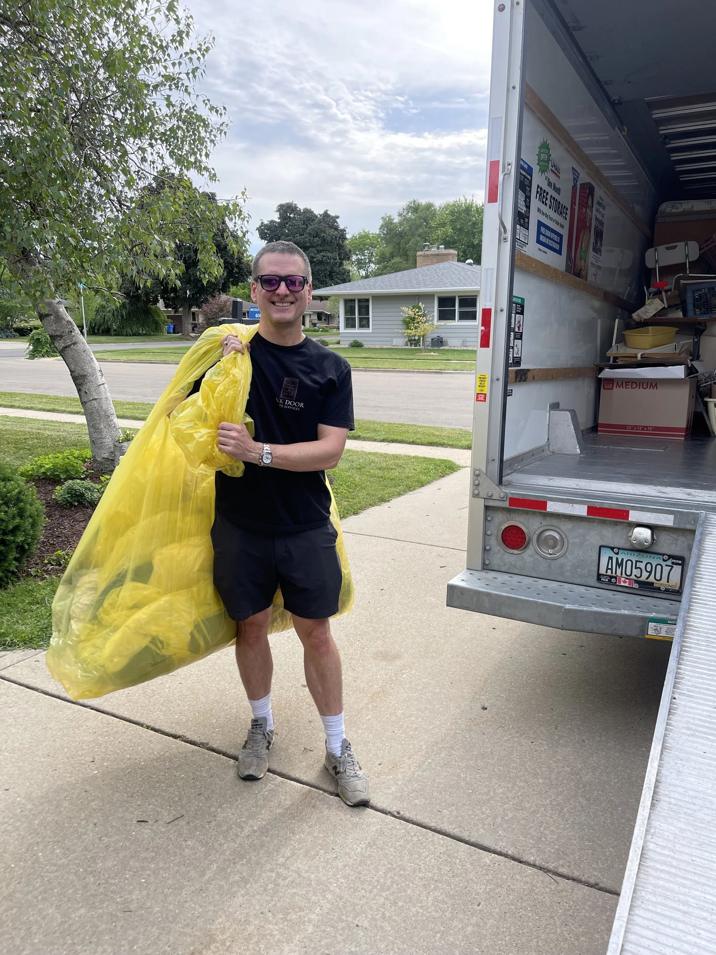 A man smiling and wearing sunglasses, a black t-shirt, black shorts, and sneakers, holding a large yellow trash bag, standing next to an empty moving truck on a suburban street with trees and houses in the background.