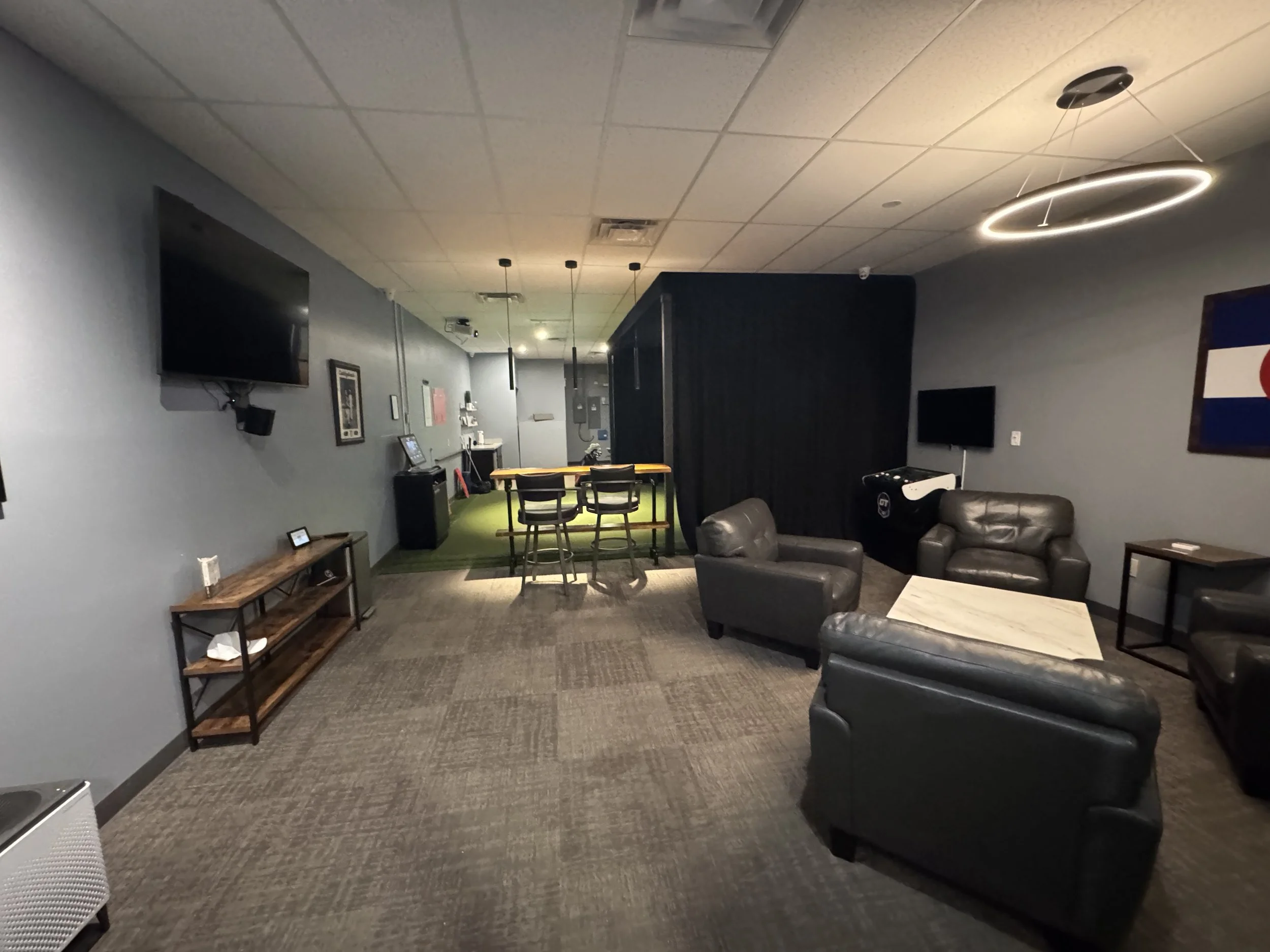Indoor lounge area with gray walls, black leather chairs, a wooden table, wall-mounted TVs, and modern circular ceiling lights.