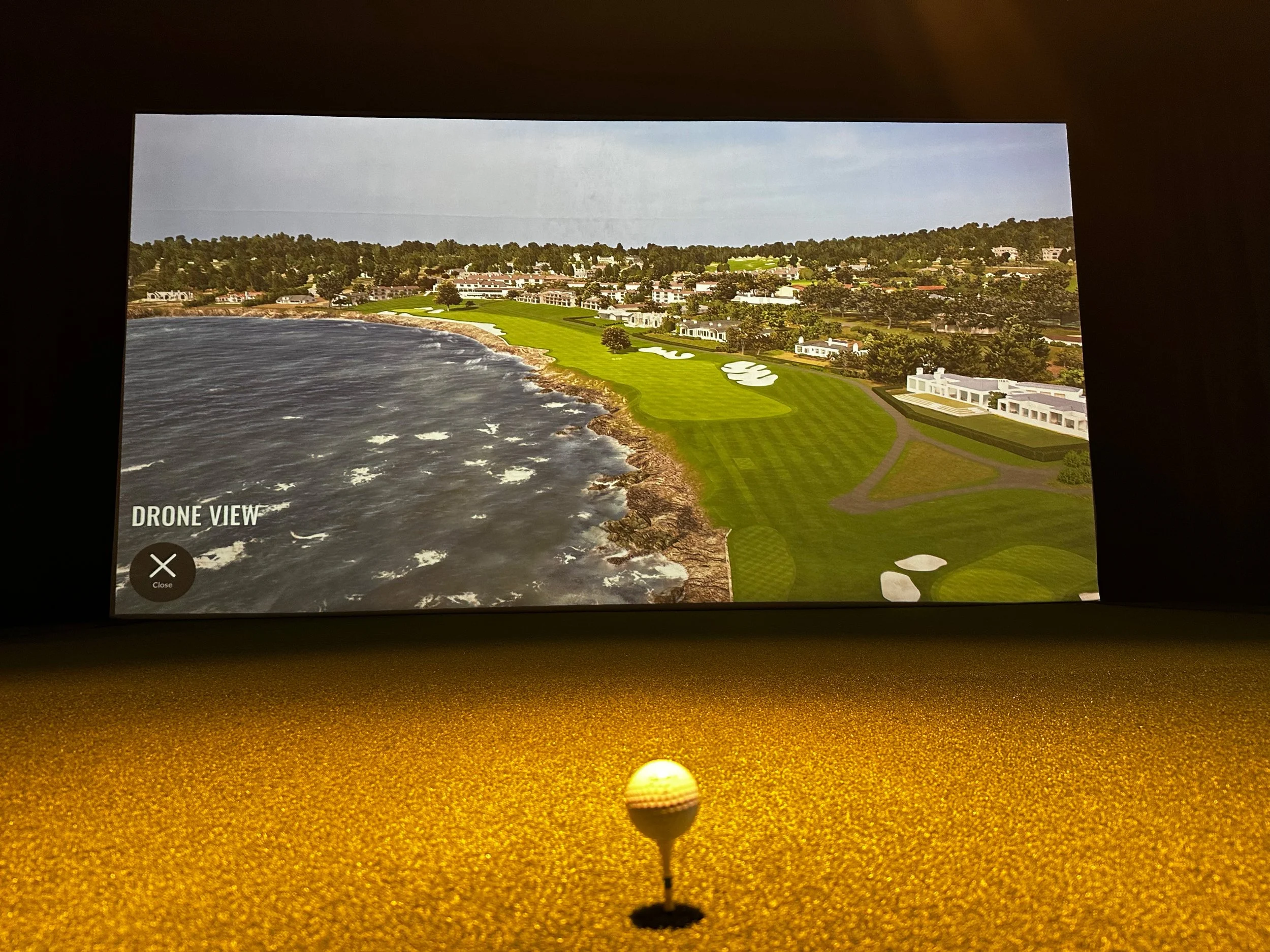 Indoor golf simulator showing a drone view of a coastal golf course with green fairways, trees, and buildings in the background, and a golf ball on a tee in the foreground.