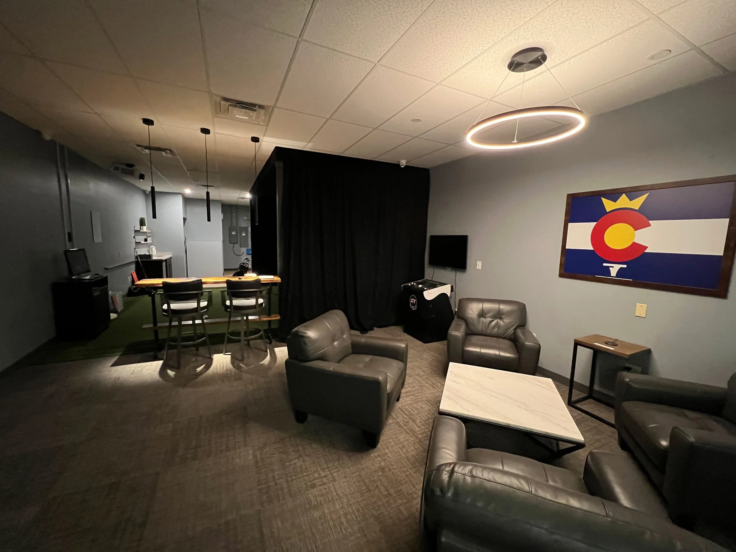 Indoor lounge area with black leather chairs and a coffee table, top right a Colorado flag painting, and a large circular ceiling light.