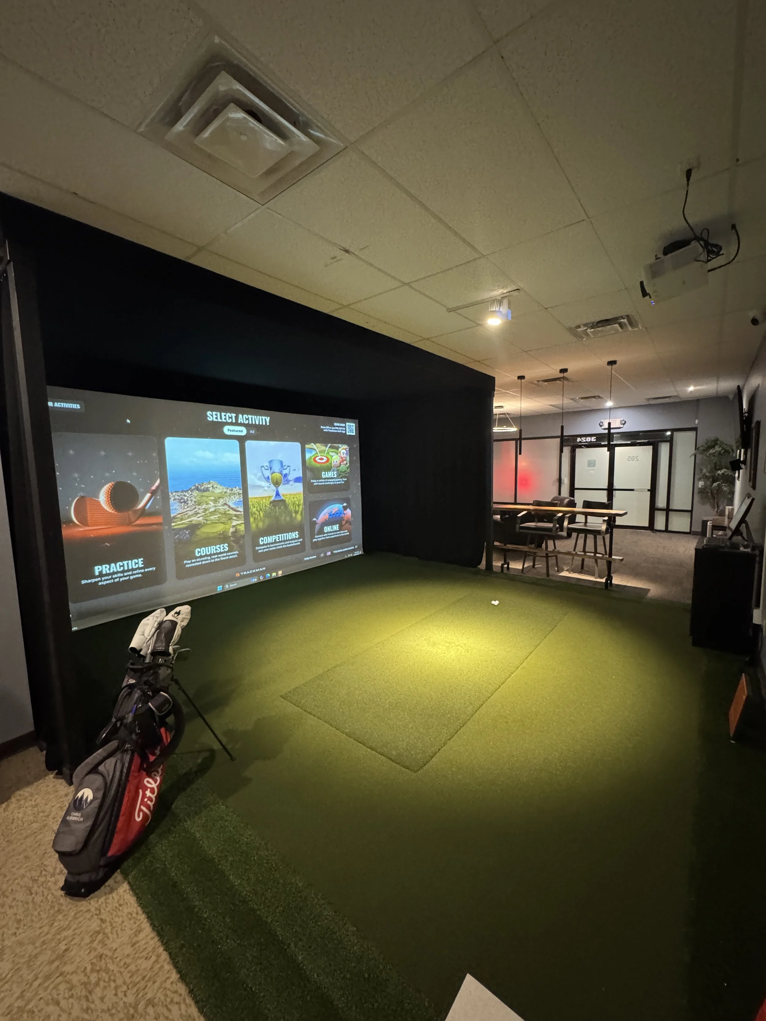 Indoor golf simulator room with a large screen displaying activity options, artificial turf area, golf bag with clubs, and seating area in background.