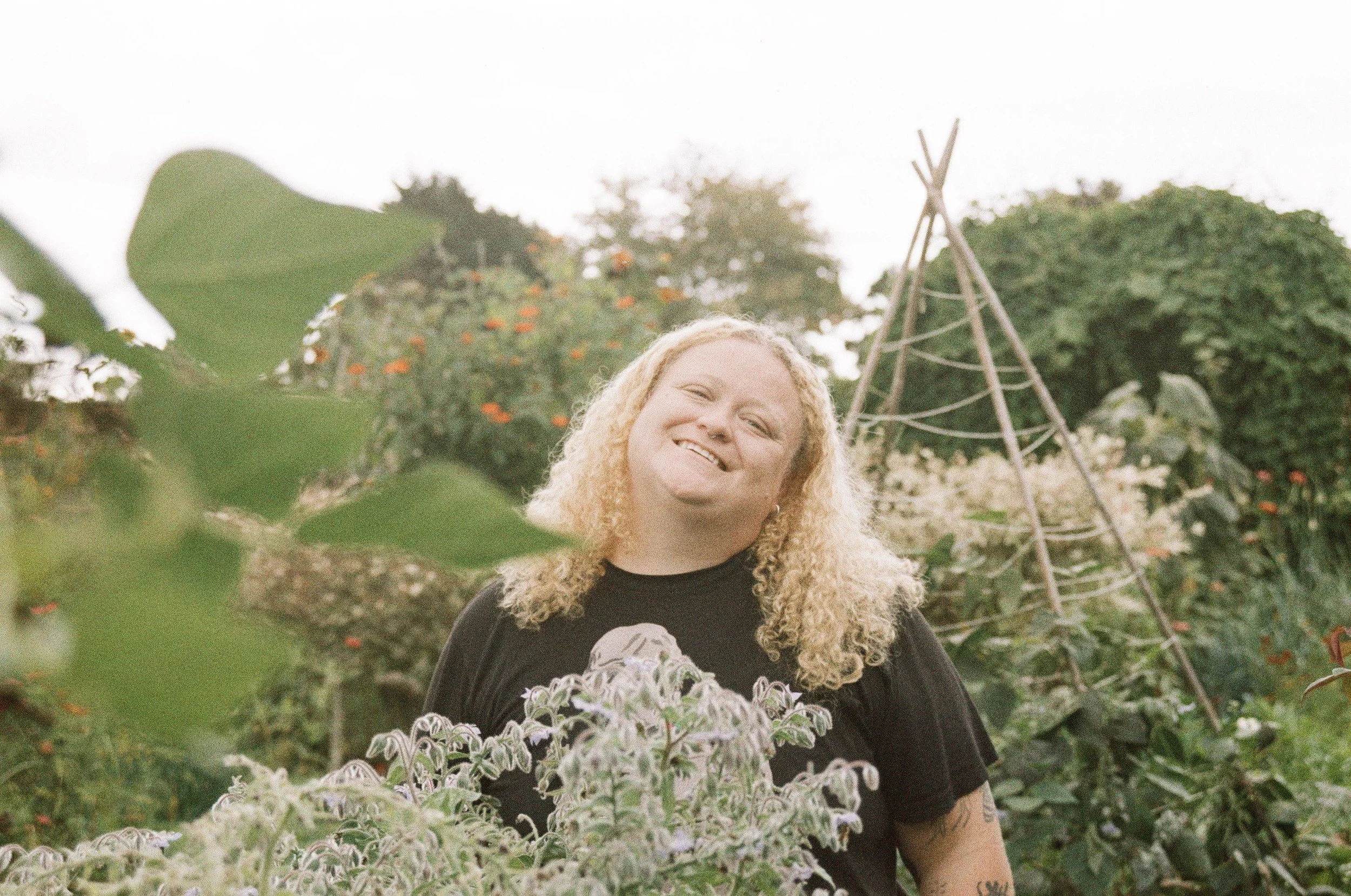 A smiling woman with curly blonde hair standing outdoors in a garden surrounded by plants, with a teepee structure in the background.