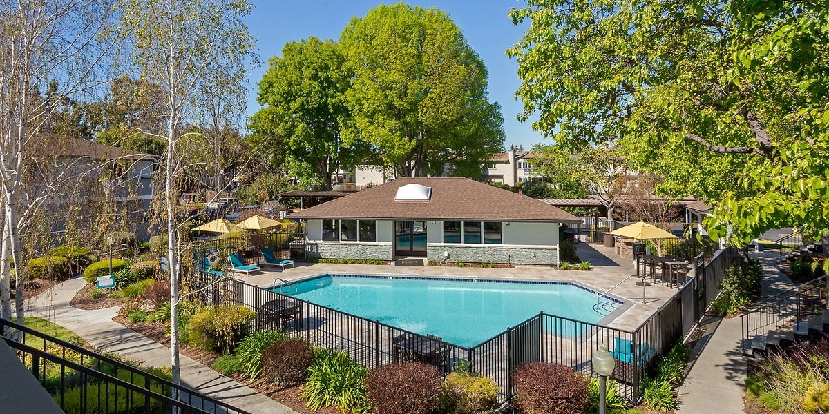 Community swimming pool surrounded by black fence, lounge chairs, umbrellas, and landscaped greenery in a residential area with trees and apartment buildings in the background.
