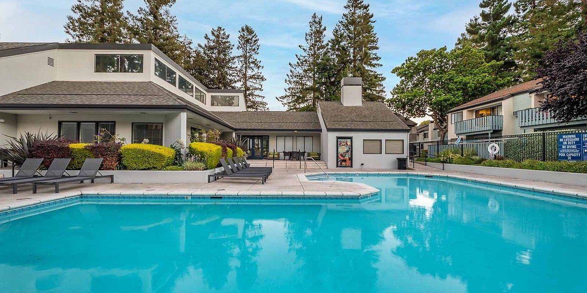 An outdoor swimming pool surrounded by lounge chairs, a landscaped area with bushes and trees, and a building with windows and balconies, in a residential or hotel complex.