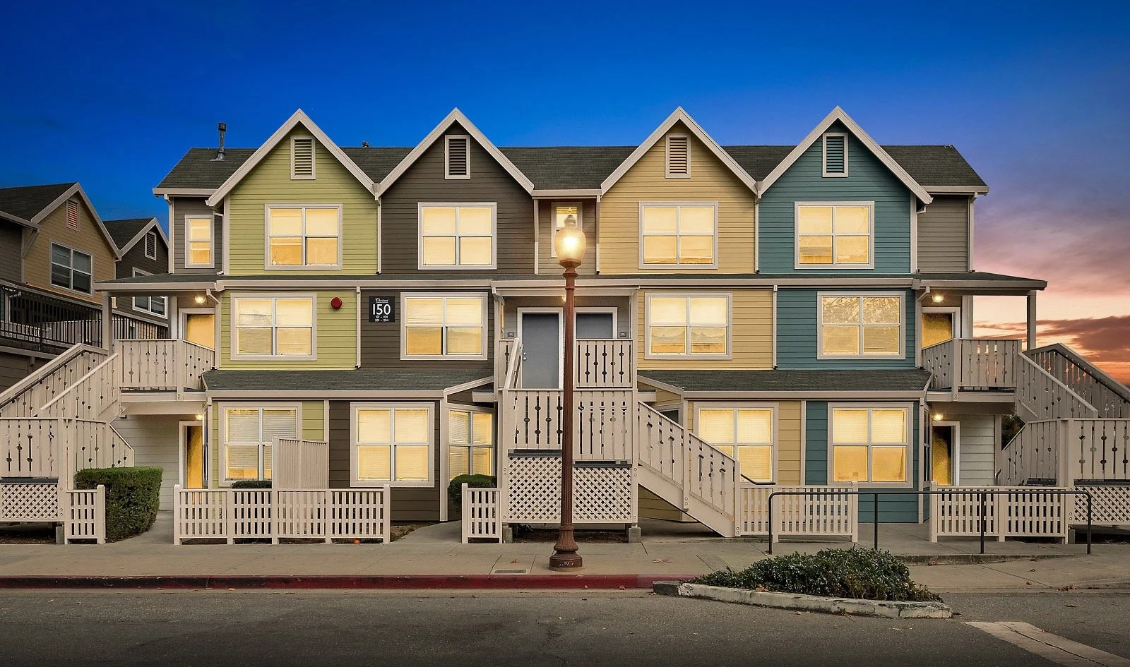 A colorful multi-story apartment building with pastel siding, illuminated windows, a central staircase, and a streetlamp in front at dusk.