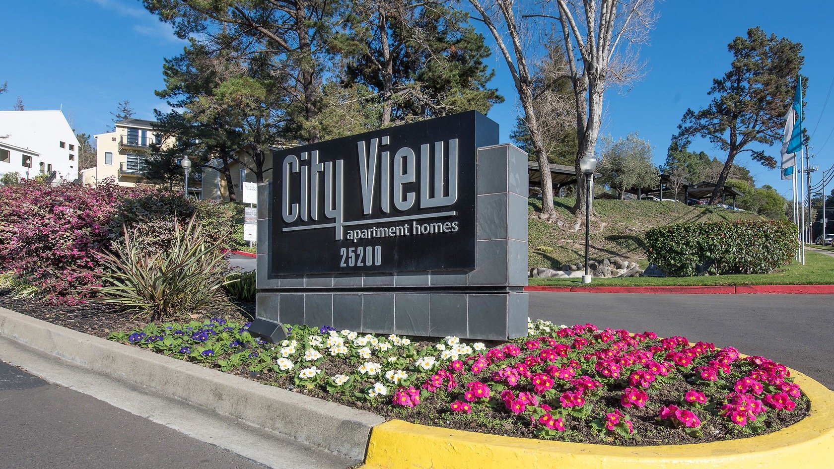 A sign for City View apartment homes with the address 25200, surrounded by pink, purple, and white flowers, and trees with buildings and blue sky in the background.