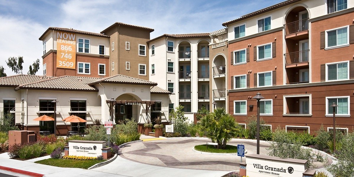 Apartment complex with modern buildings, a leasing office labeled 'Leasing' at the entrance, outdoor seating with umbrellas, landscaping with plants and small trees, and signs indicating apartment homes for rent in the Villa Granada community.